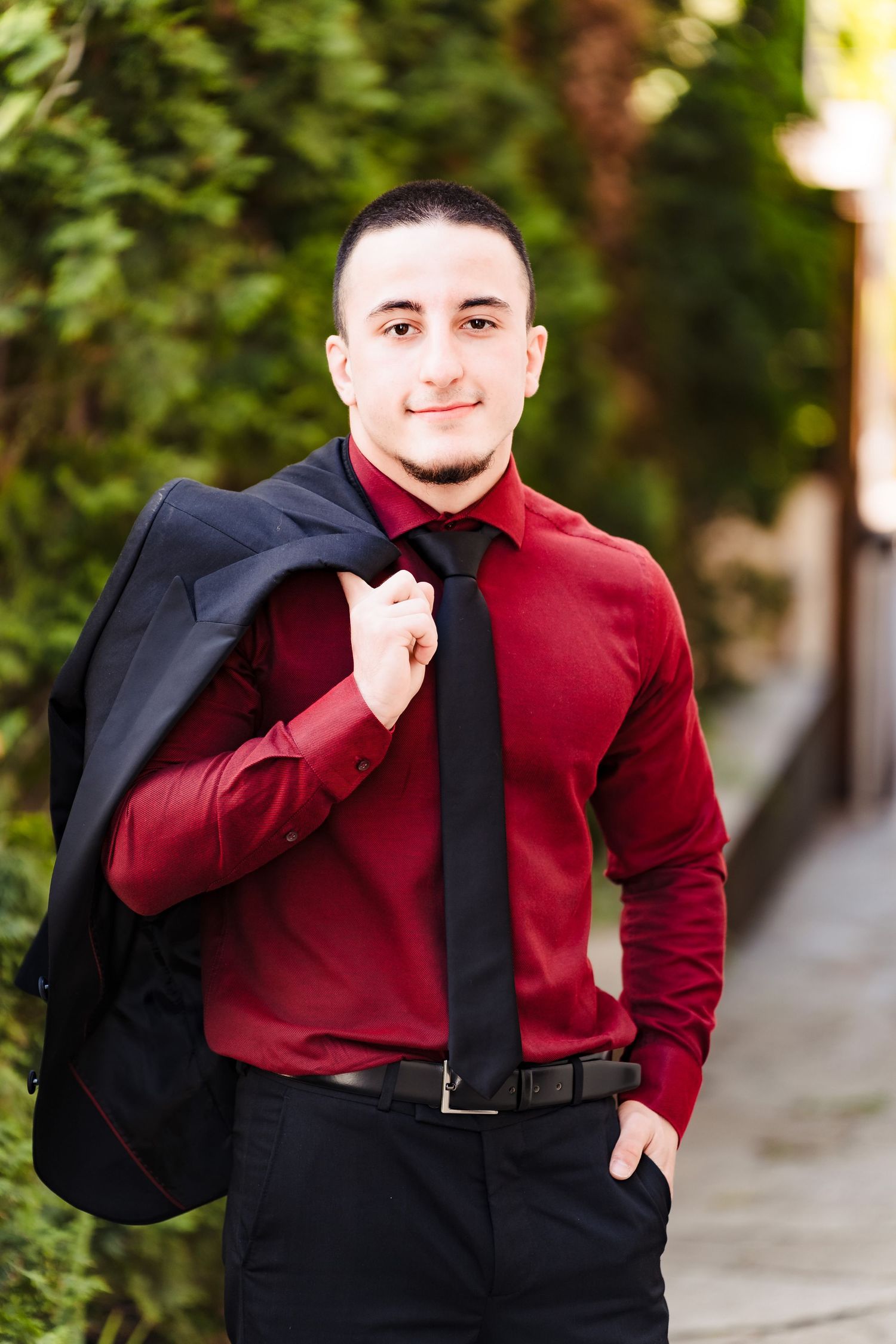 Smiling young man in a burgundy dress shirt and black tie holding a dark suit jacket over his shoulder outdoors.