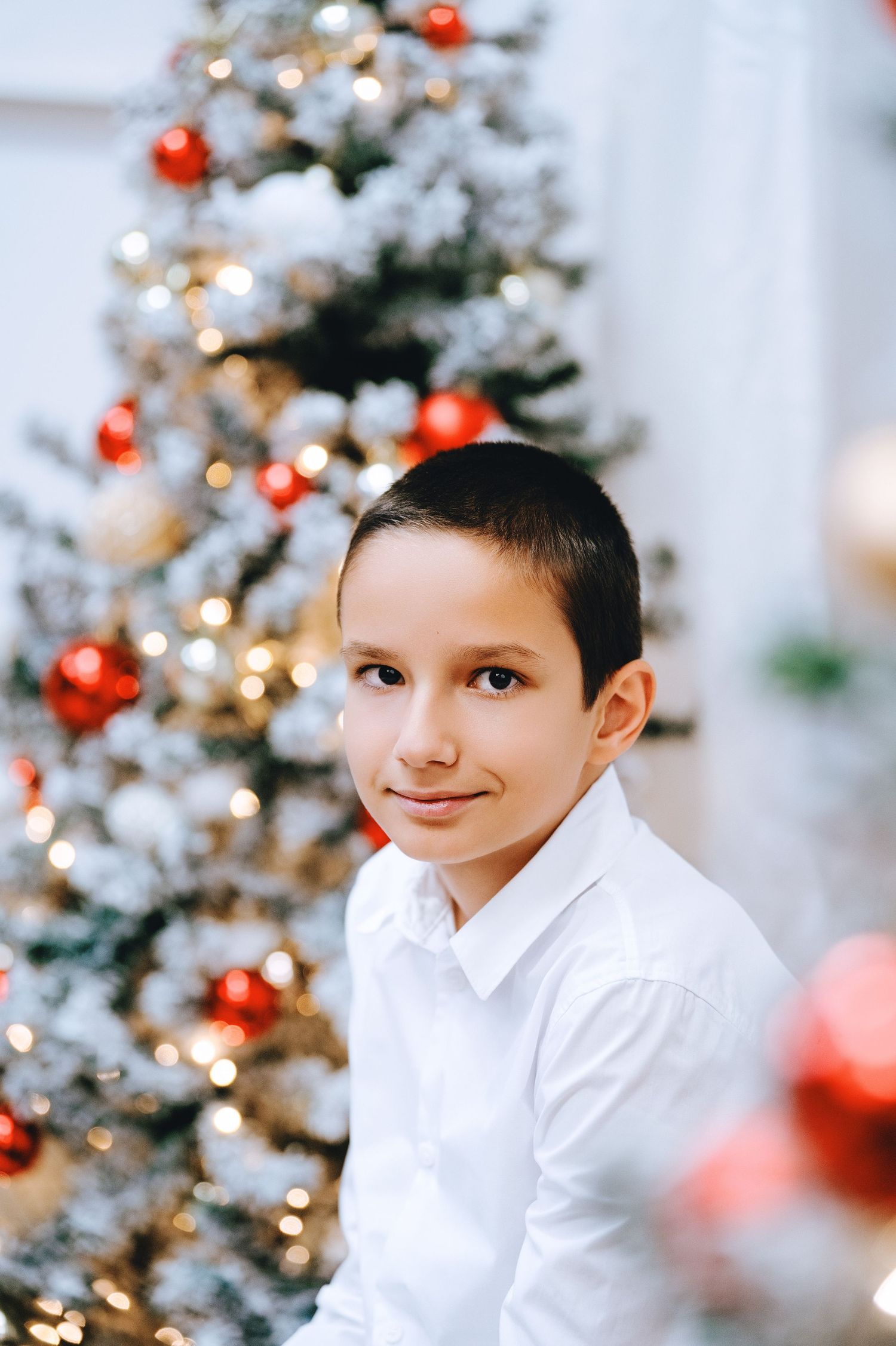 Smiling boy in a white button-up shirt stands in front of a flocked Christmas tree decorated with red ornaments and warm glowing lights.