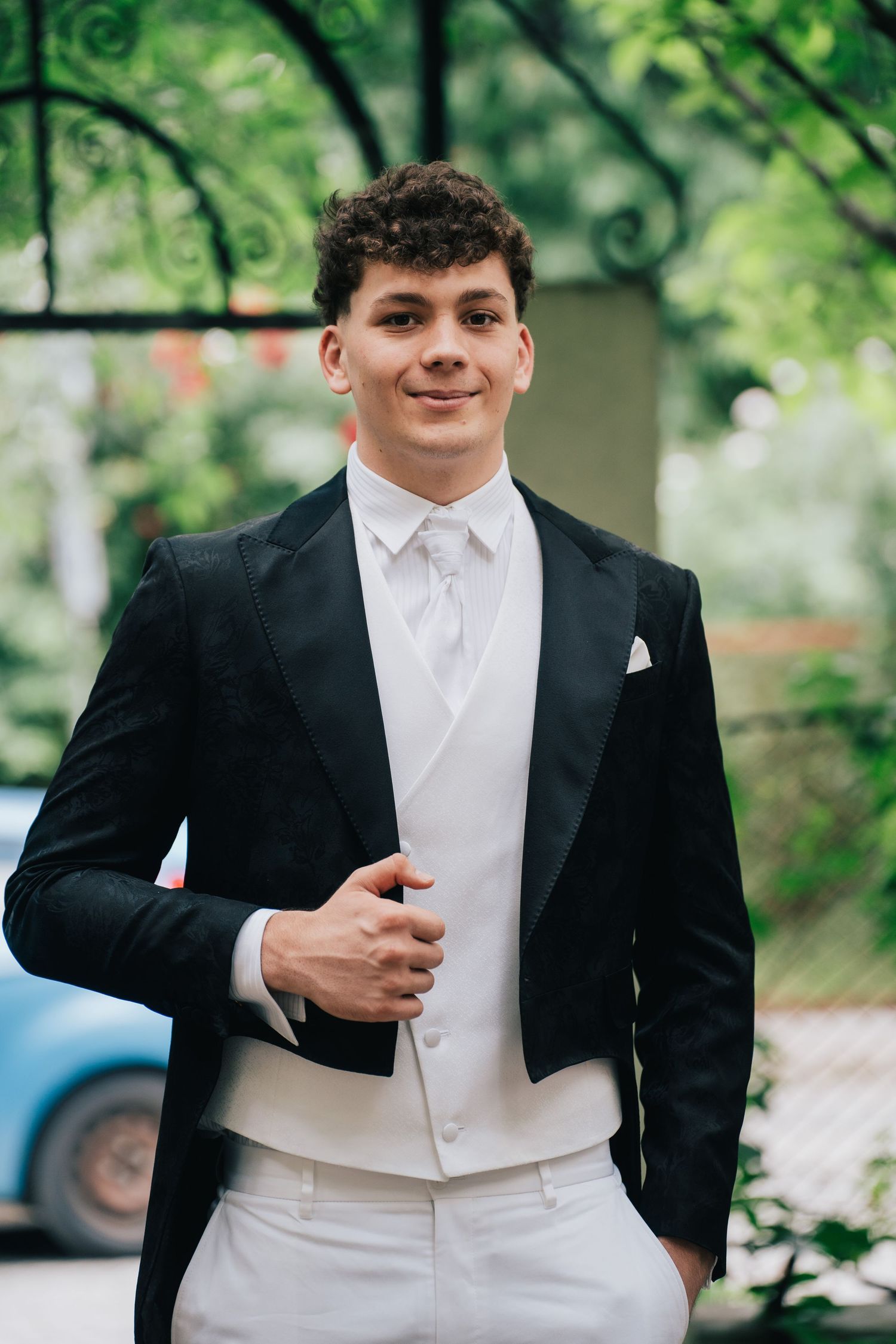 Smiling young man in a black tailcoat, white vest and light trousers stands outdoors with a hand in his pocket.