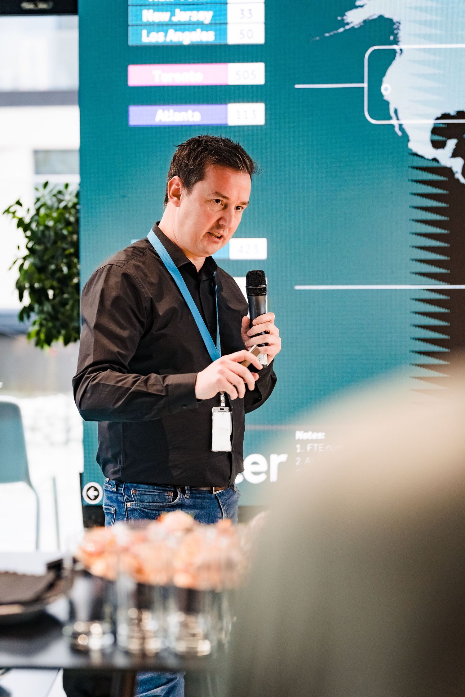 Man with a lanyard speaks into a microphone at a company event, with a presentation screen and snacks nearby.
