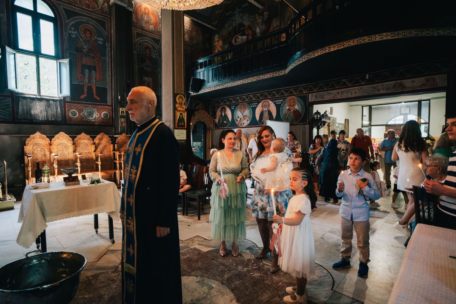 Orthodox priest in black vestments stands beside a table with candles as families and children gather inside an ornate frescoed church.