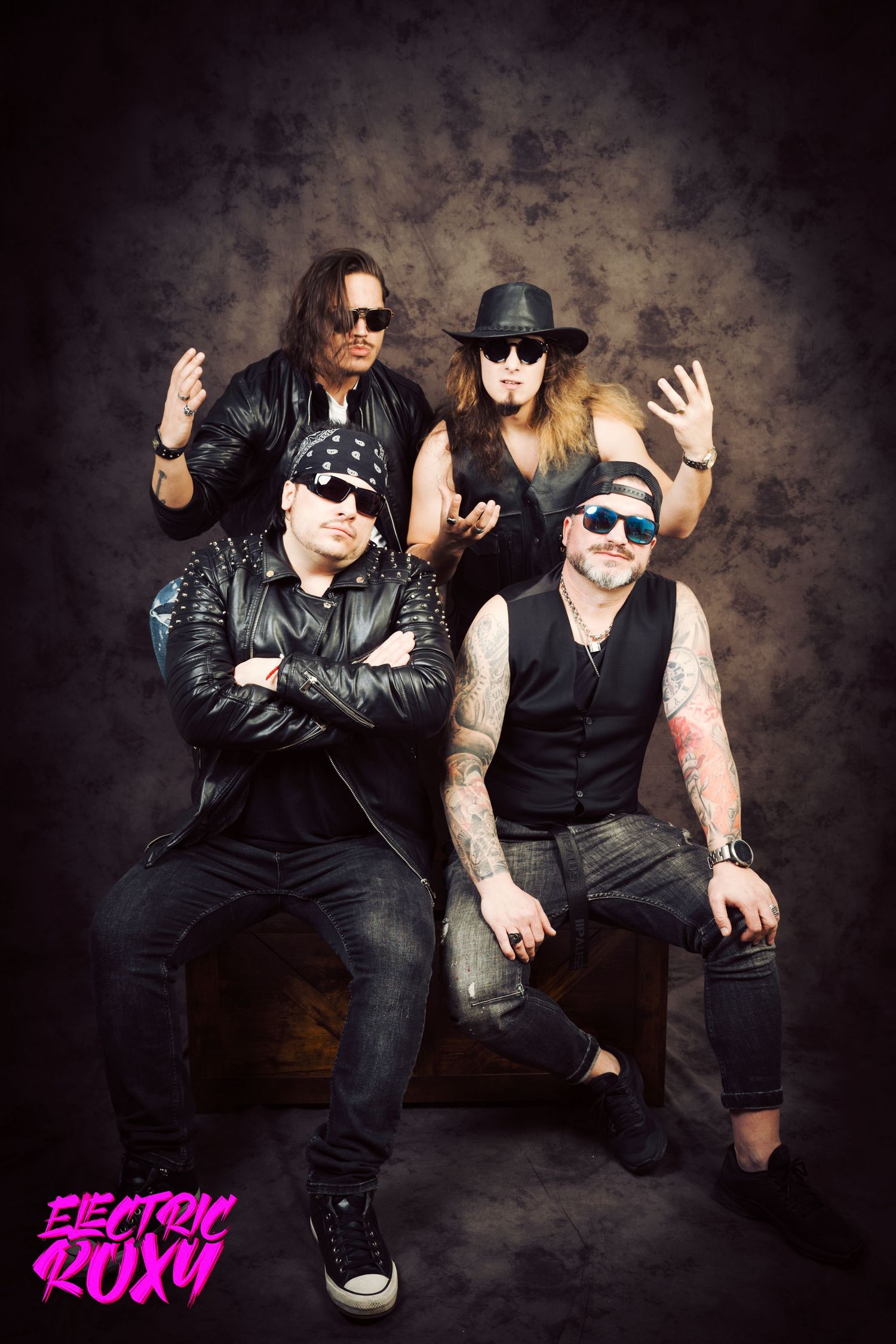 Electric Roxy rock band poses in a studio, two members seated on a wooden crate and two standing behind, wearing leather and sunglasses.