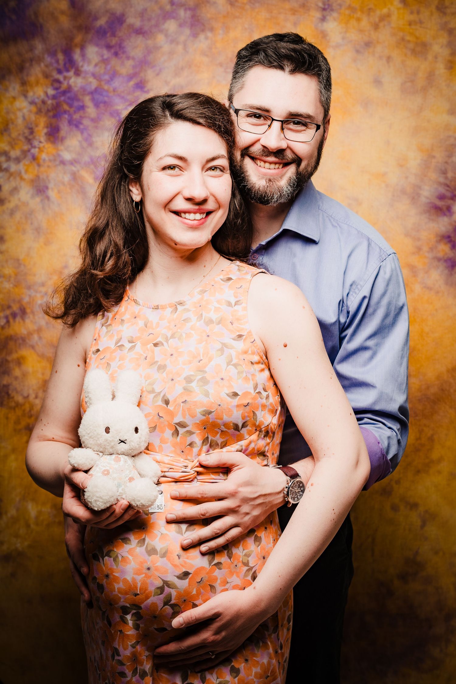 Pregnant woman holding a small white bunny plush while her smiling bearded partner hugs her from behind in a studio portrait.