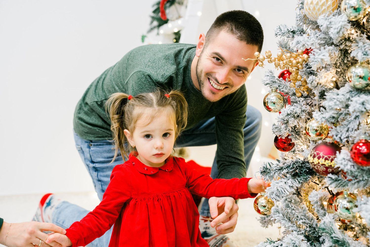 Smiling father and toddler girl in a red dress decorating a Christmas tree with shiny red and gold ornaments.