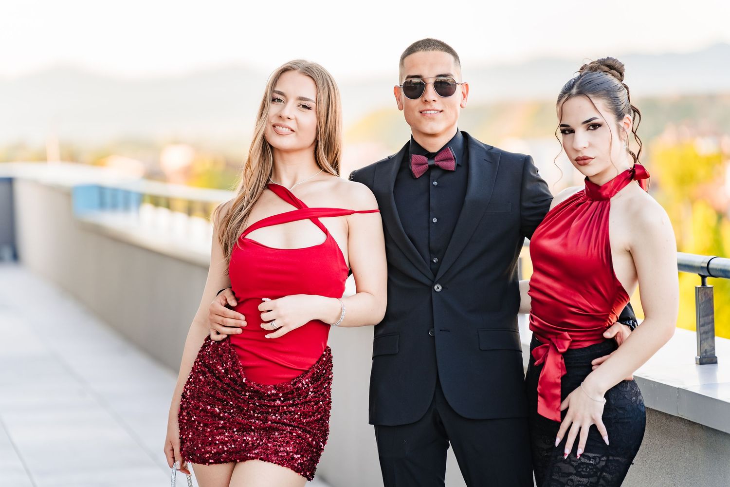 Young man in a black suit and bow tie posing with two women in red outfits on a terrace overlooking green hills.