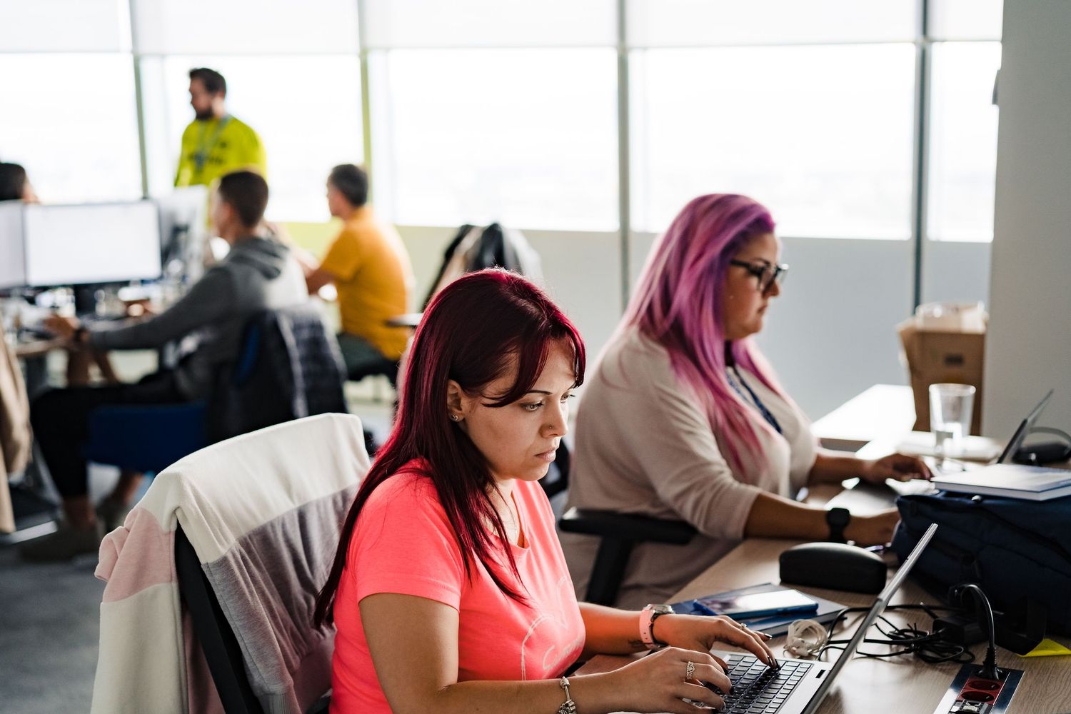 Colleagues work on laptops and computers in an open-plan office, including a woman with pink hair and glasses typing in the foreground.