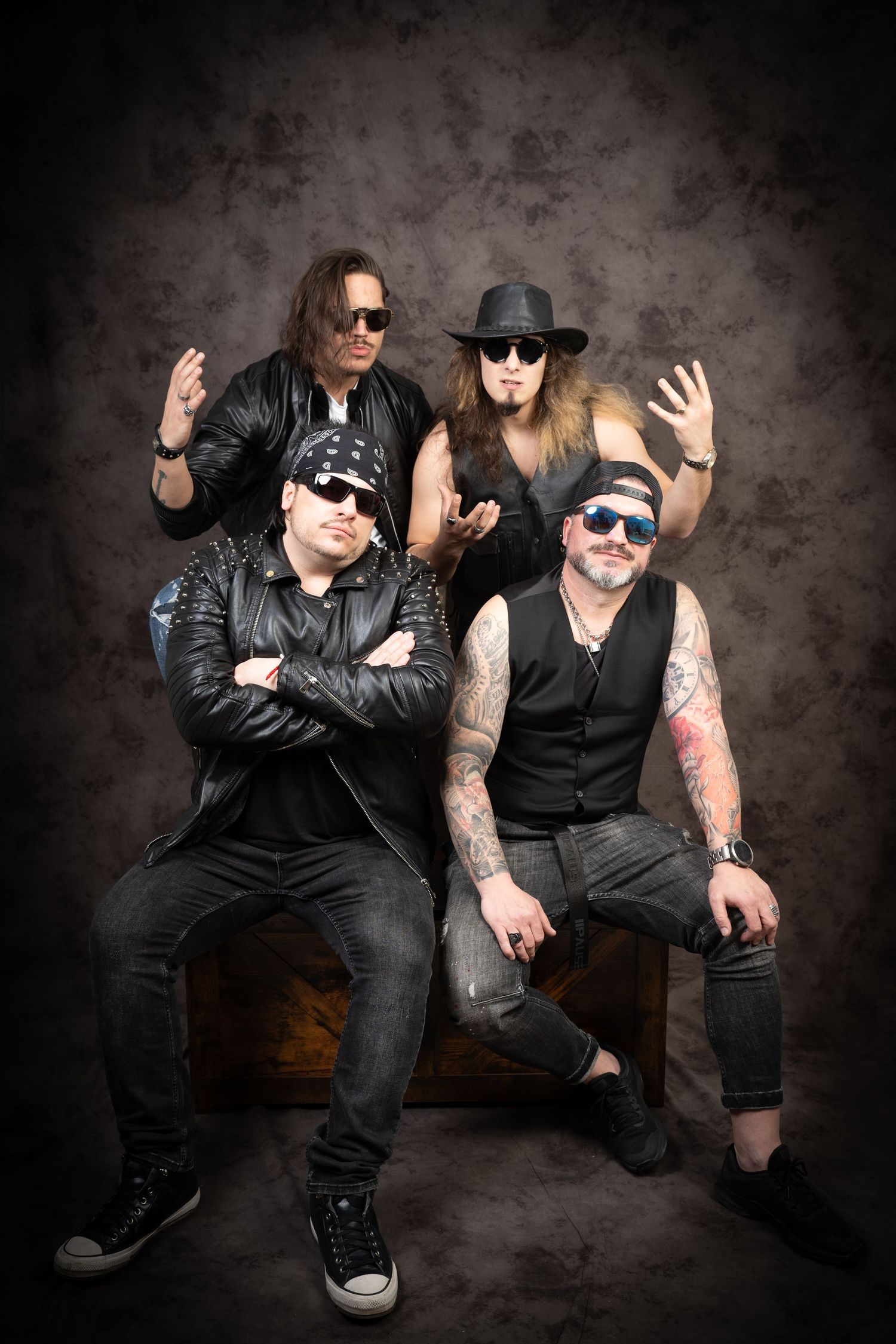 Four men in black leather jackets, sunglasses and hats pose in a studio against a dark textured backdrop, two seated and two standing.