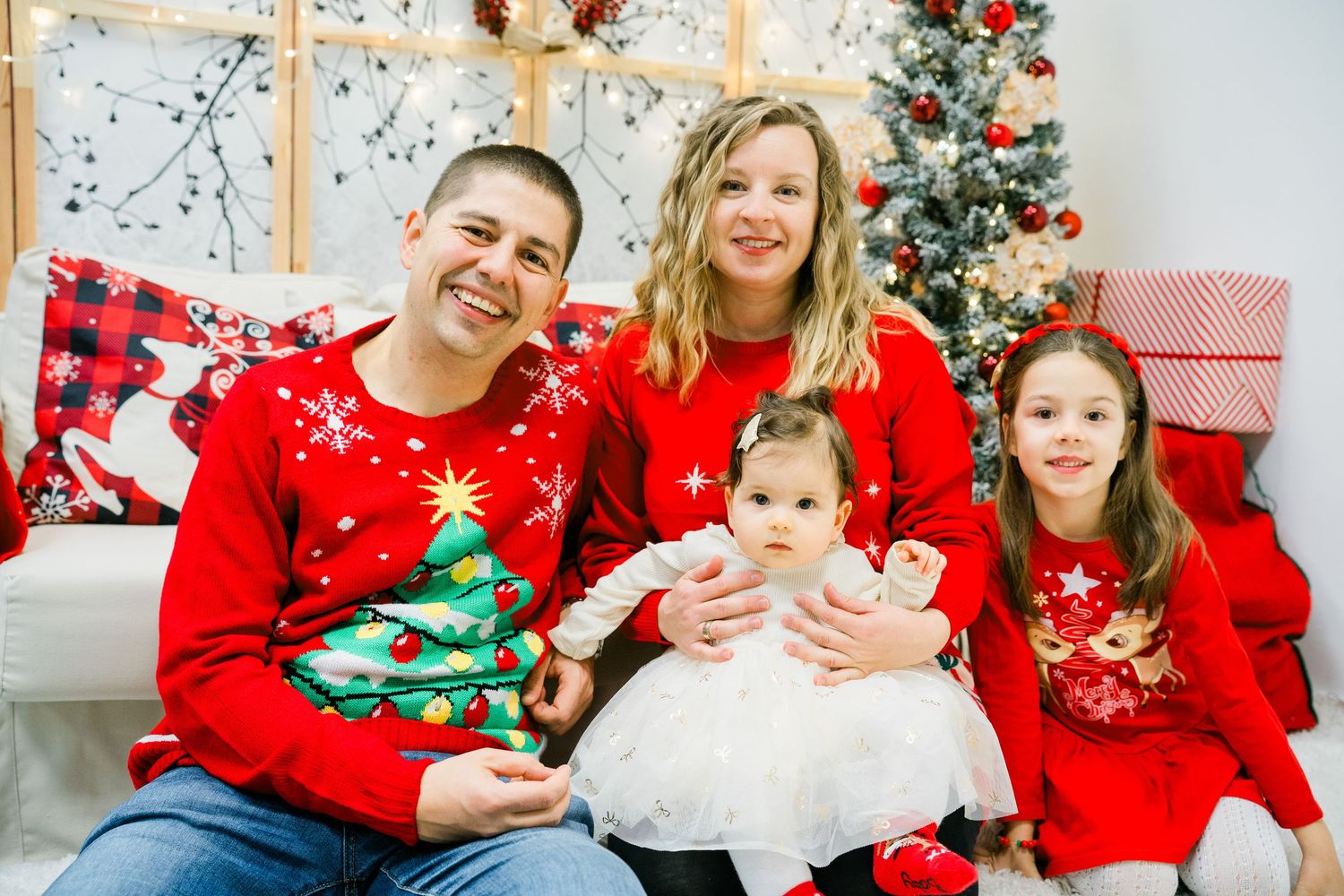 Smiling family in matching red Christmas sweaters poses with a baby and young girl beside a decorated holiday tree.