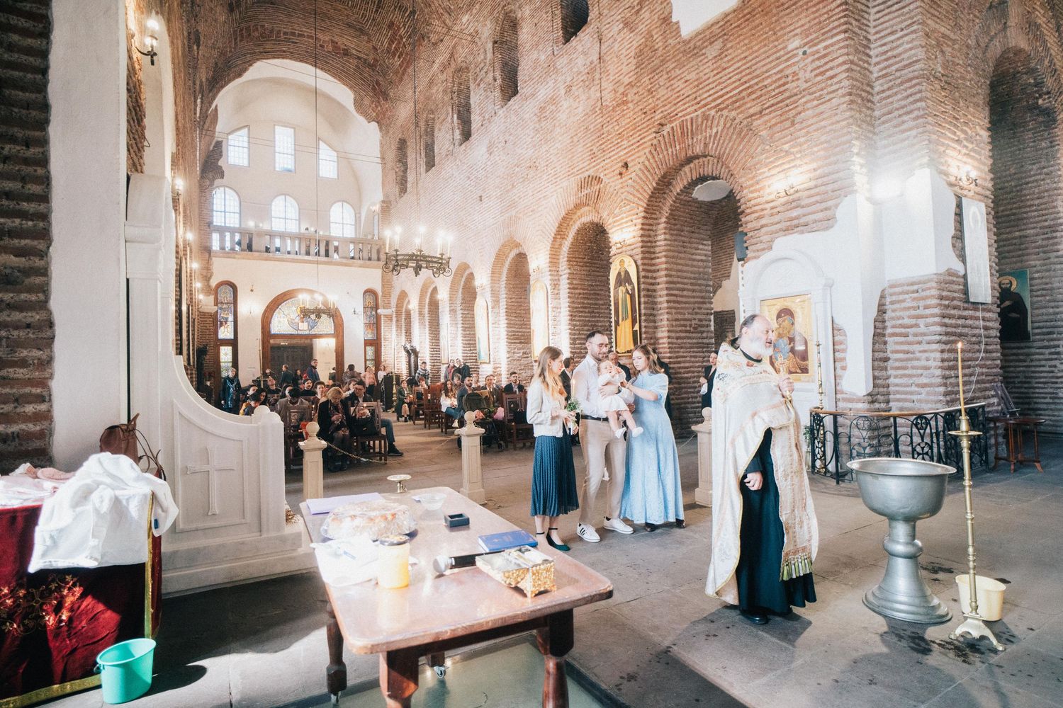 Family and guests gather around a priest in vestments in a brick church for an Orthodox baptism beside the baptismal font.