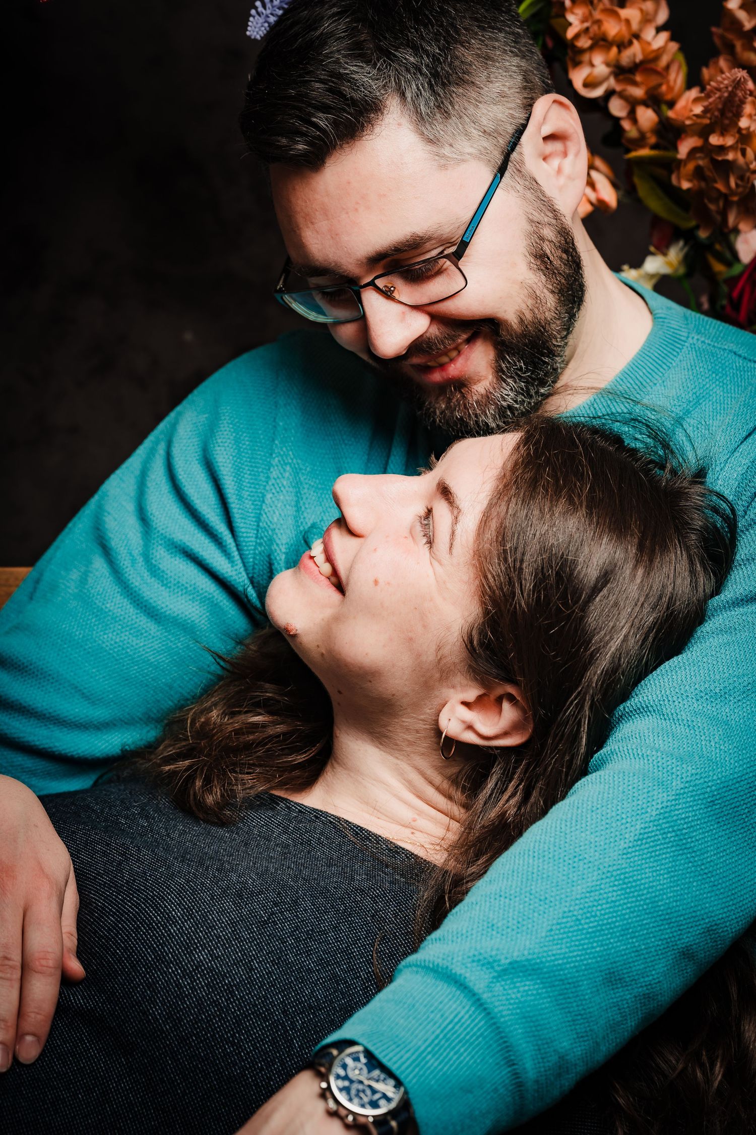 A bearded man in glasses and a teal sweater smiles lovingly down at a woman resting against him in a romantic couple portrait.