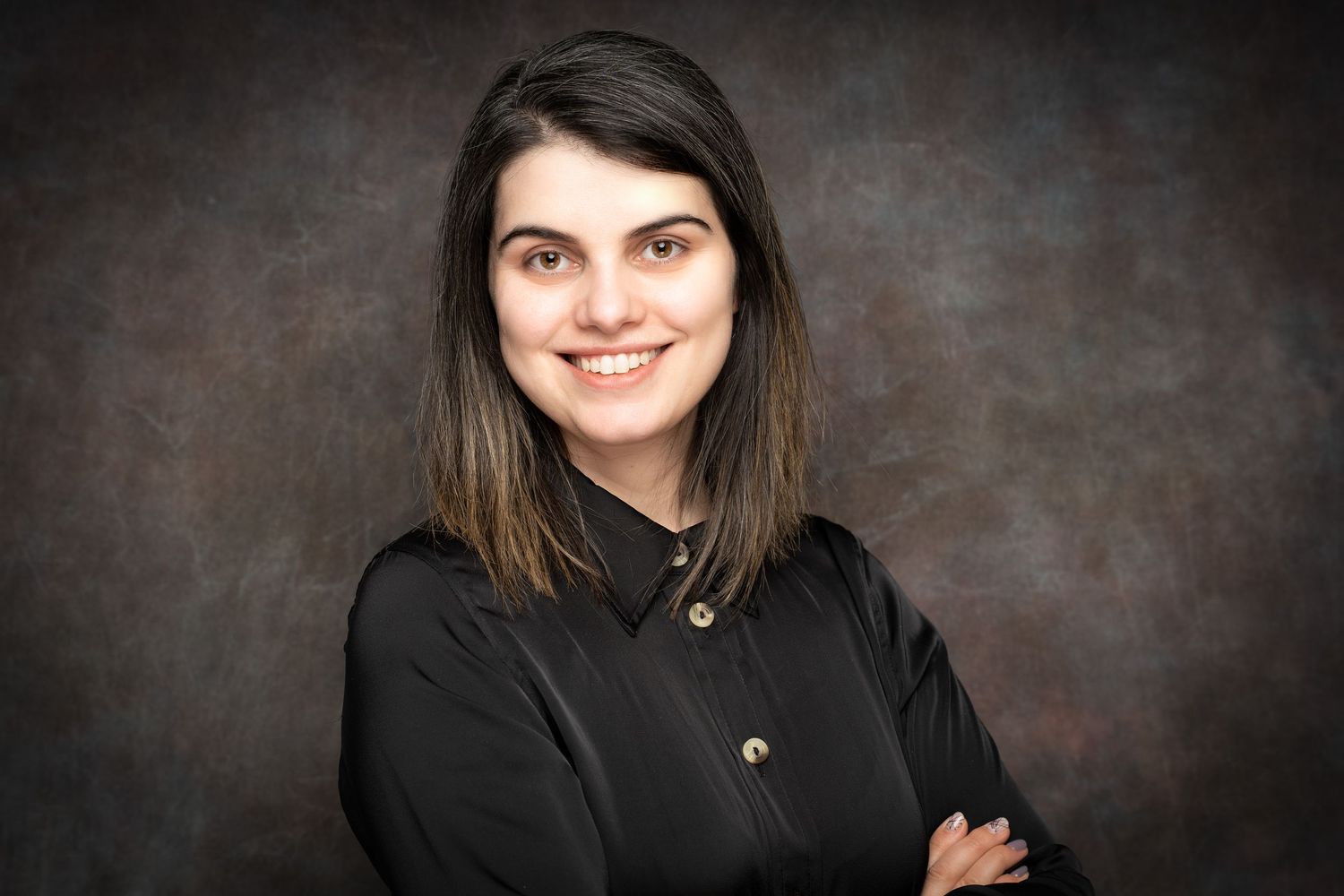 A smiling young woman with long brown hair and a black button-up shirt poses with crossed arms against a dark textured studio backdrop.