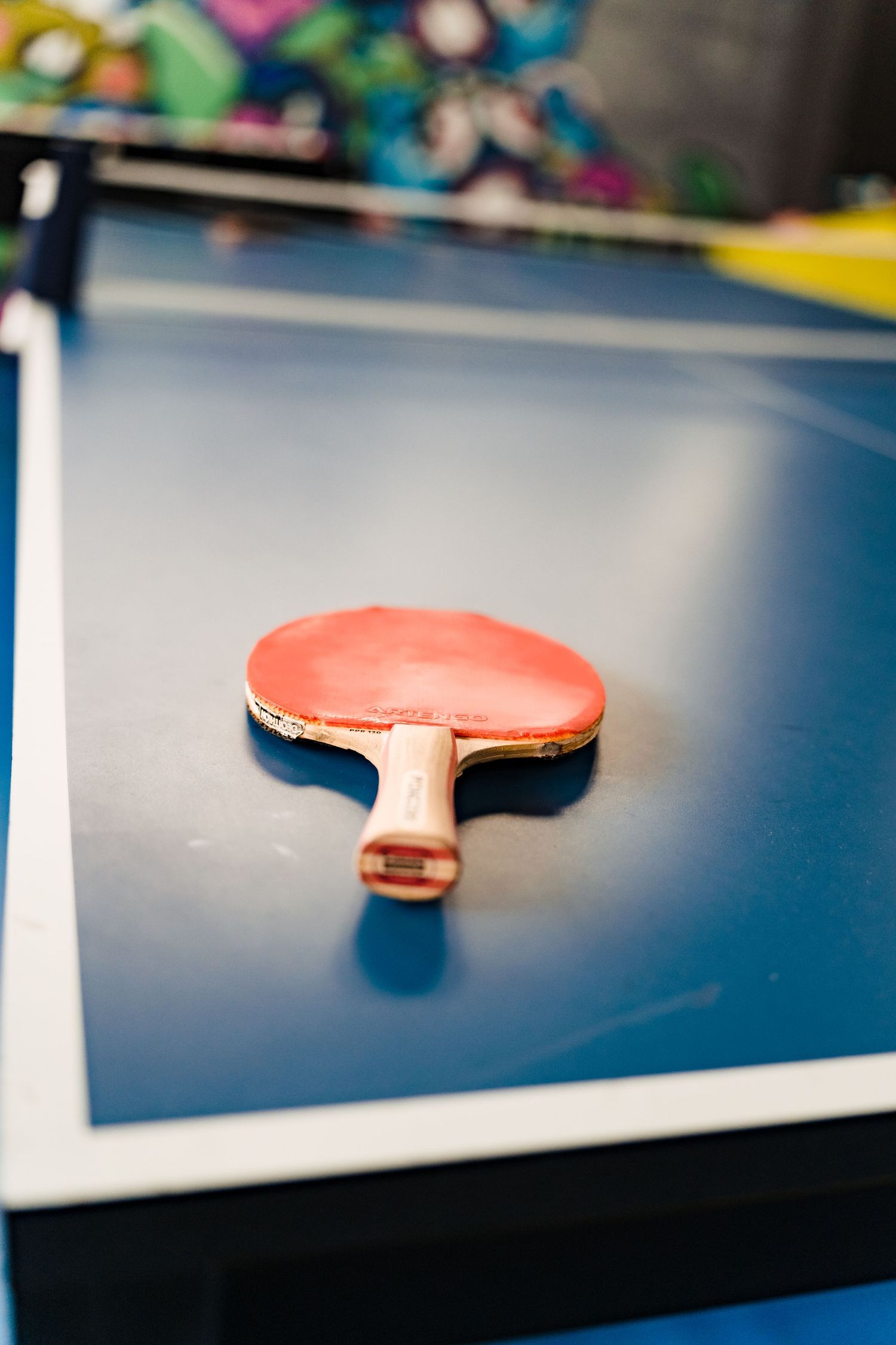 Red table tennis paddle rests on a blue ping pong table in a colorful office recreation area.