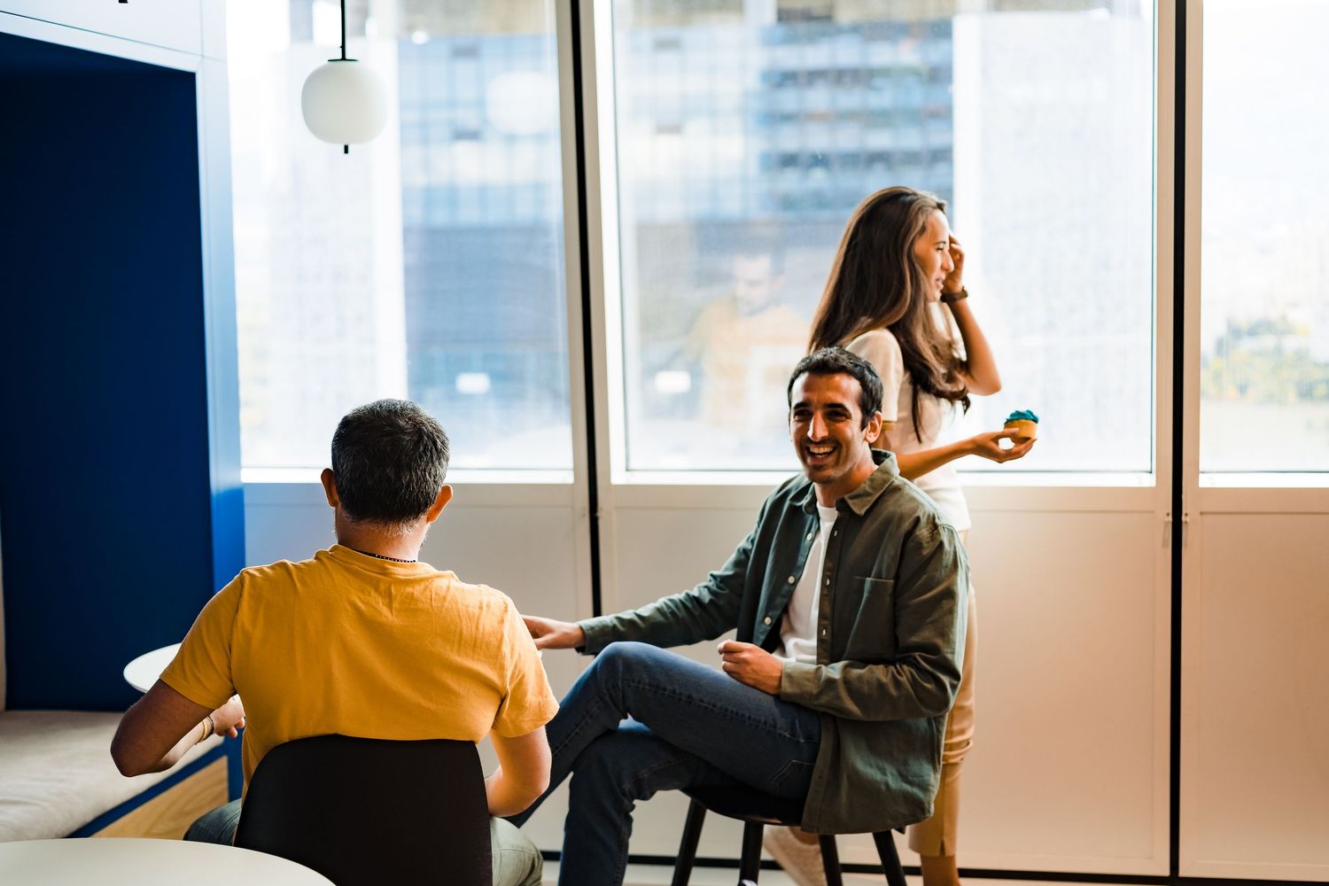 A smiling man sits on a stool talking with a standing woman and a seated colleague near large windows in a modern office.