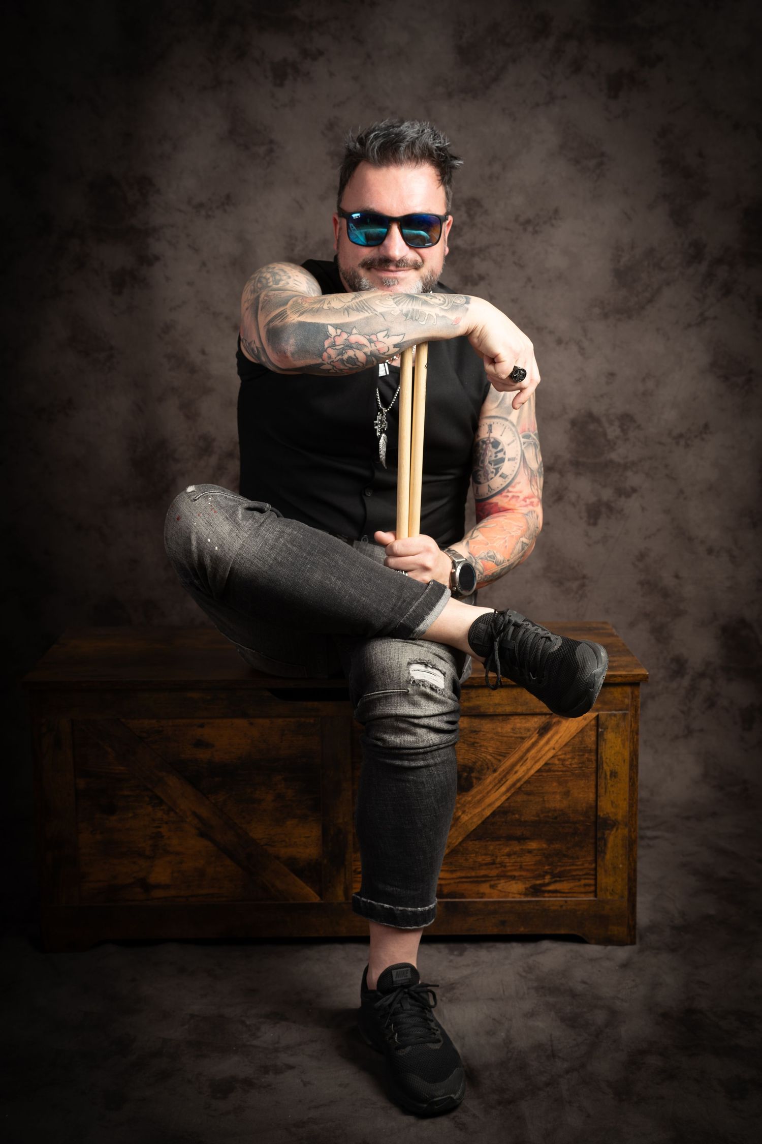 Tattooed man in blue sunglasses and a black vest sits on a wooden chest holding drumsticks in a studio portrait.