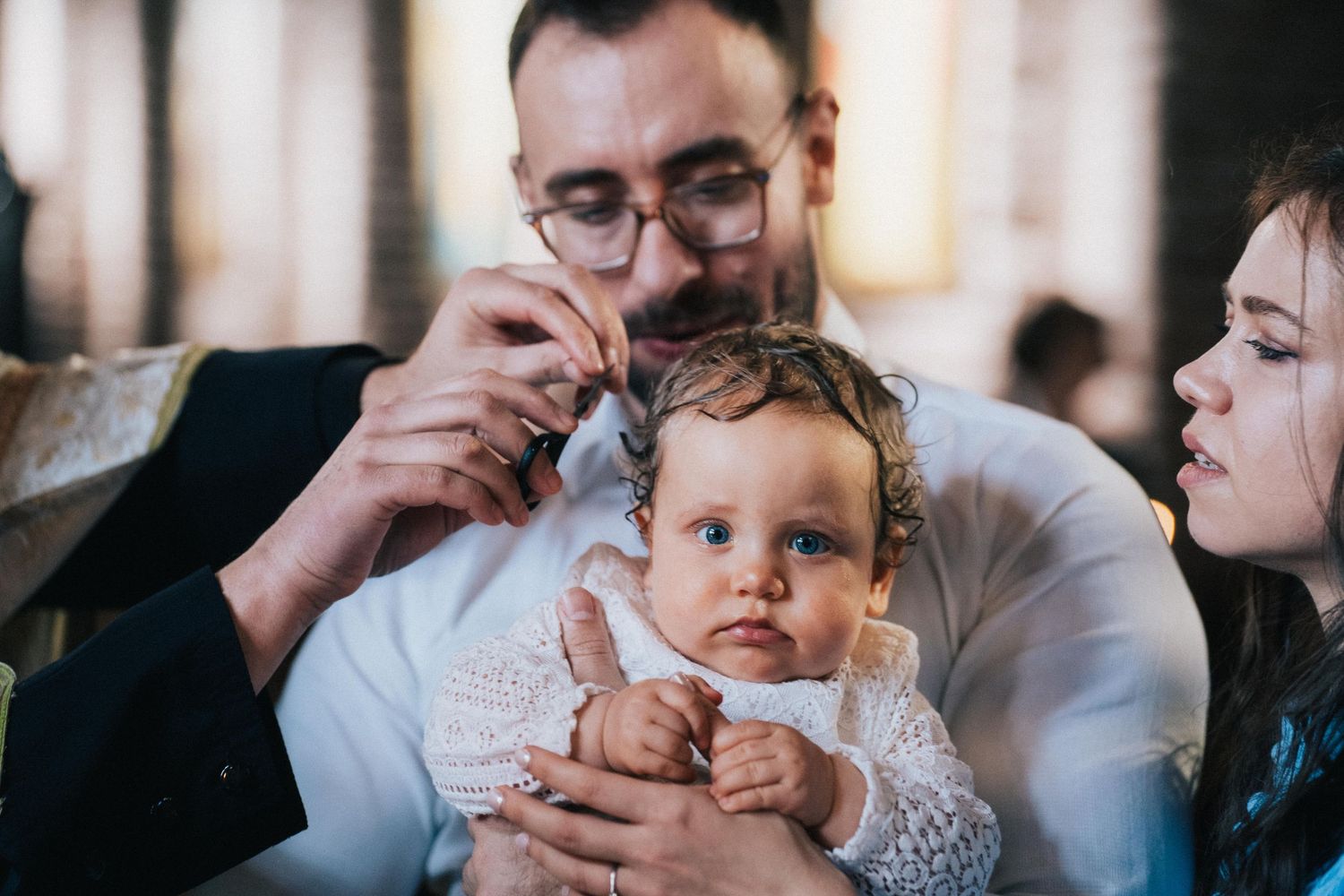 A priest anoints a baby's forehead during an Orthodox baptism as the father holds the child and the mother watches closely.