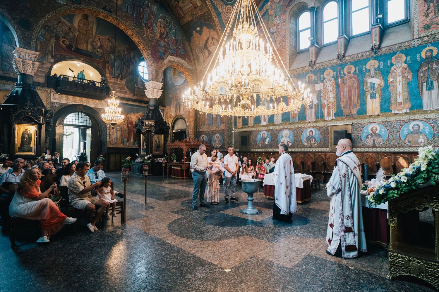 Priests and family gather around a baptismal font beneath a glowing chandelier and iconic frescoes in a traditional Orthodox church.