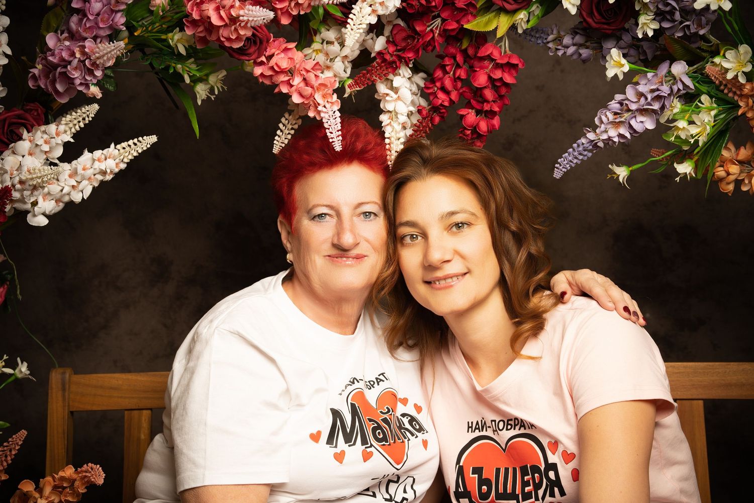 A red-haired woman and a young woman in matching white shirts embrace and smile beneath a hanging floral arrangement.