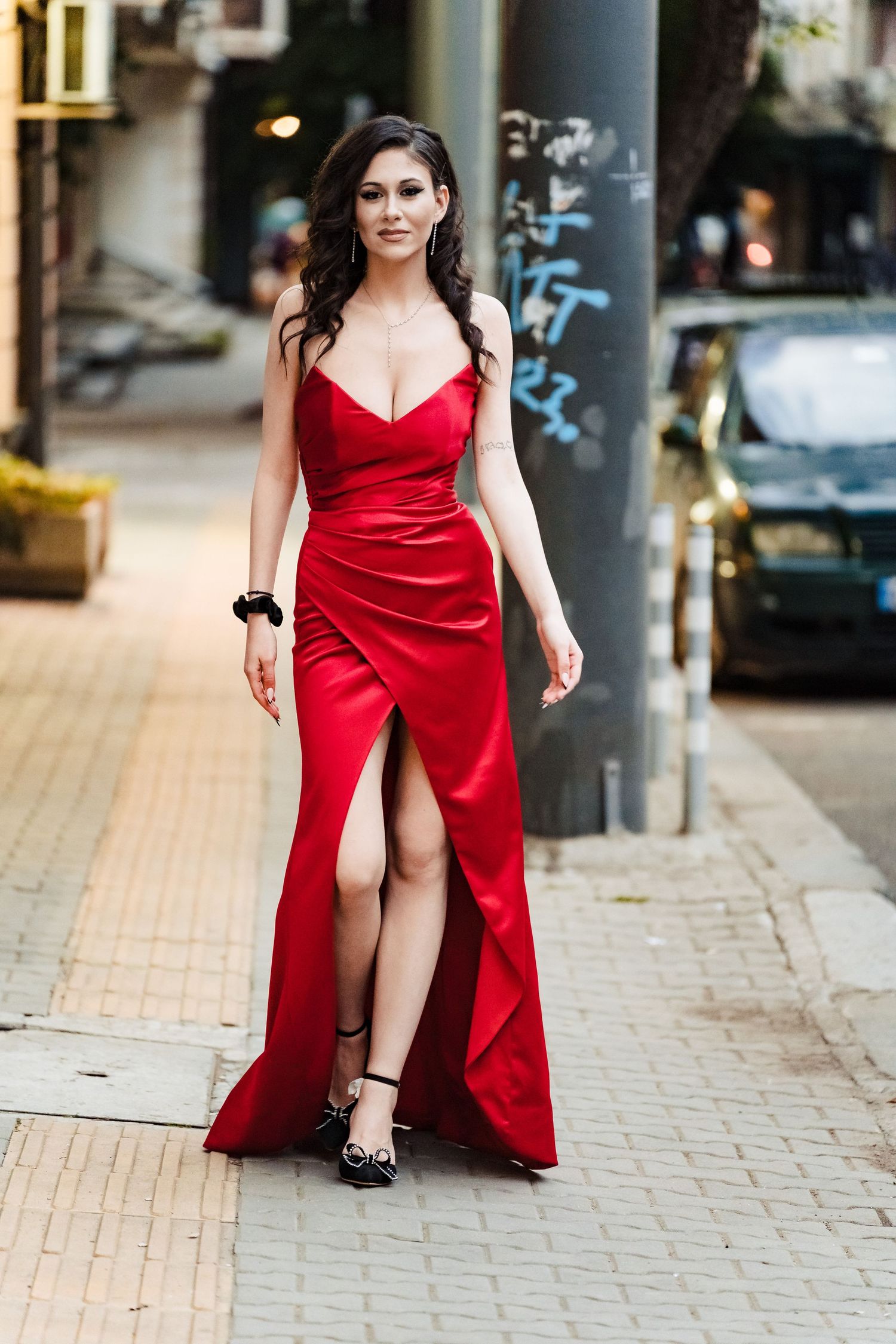 Woman in a red satin evening dress with a front slit and a black wristband poses on a cobbled city street.
