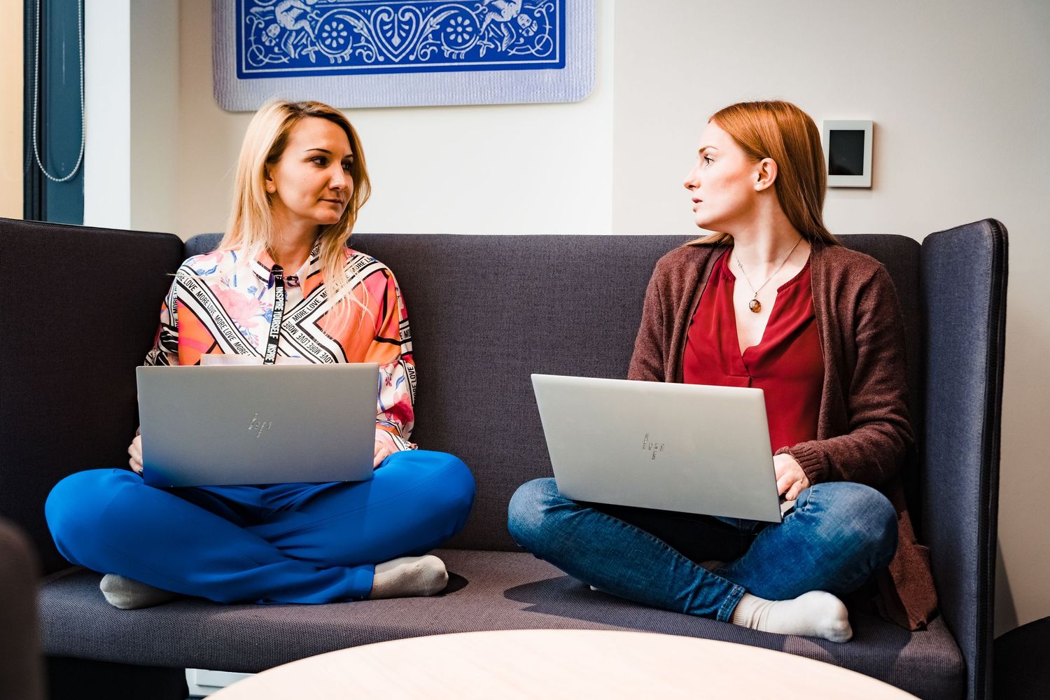 Two women sit cross-legged on a gray sofa with laptops, having a conversation in a bright, modern office lounge.