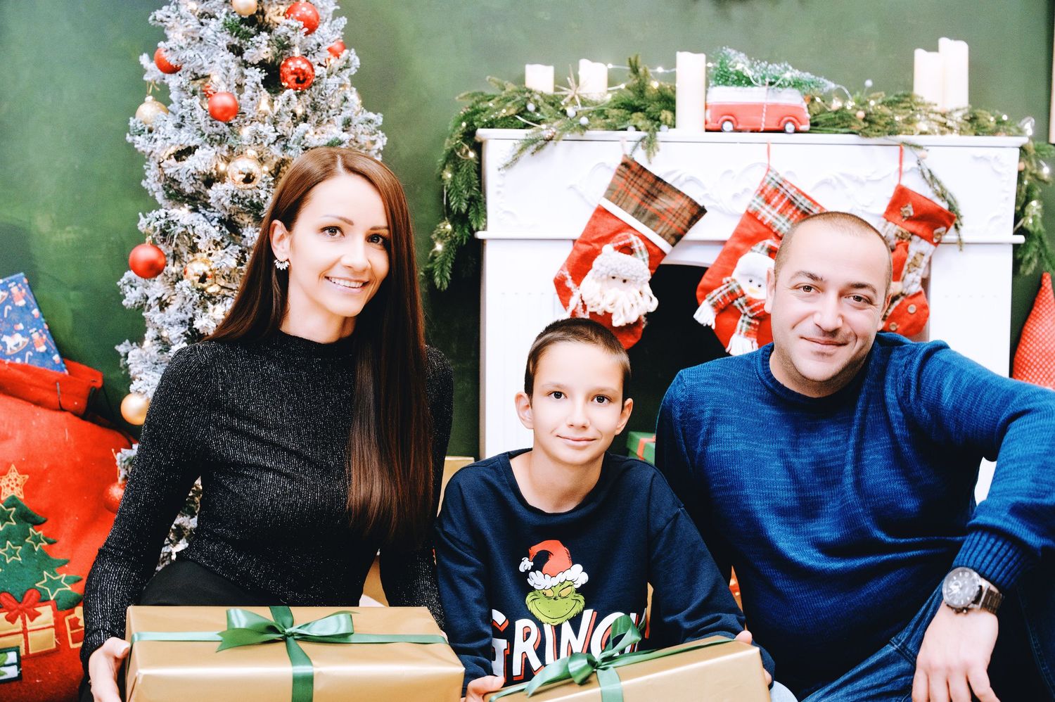 Smiling family of three poses by a decorated Christmas tree and fireplace with stockings, with wrapped gifts in the foreground.
