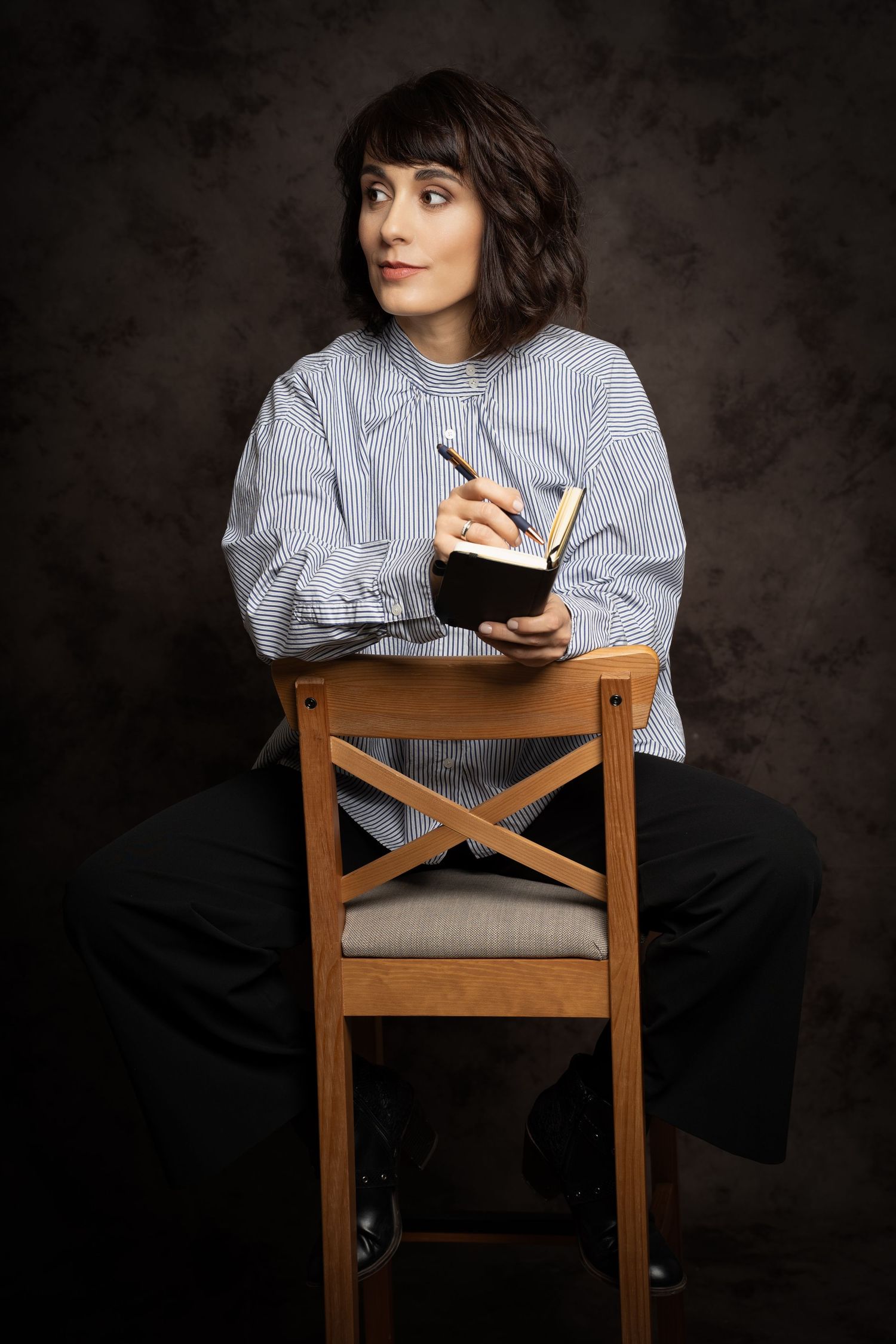 Woman in a striped blouse sits on a wooden chair holding a pen and small notebook in a moody studio portrait.