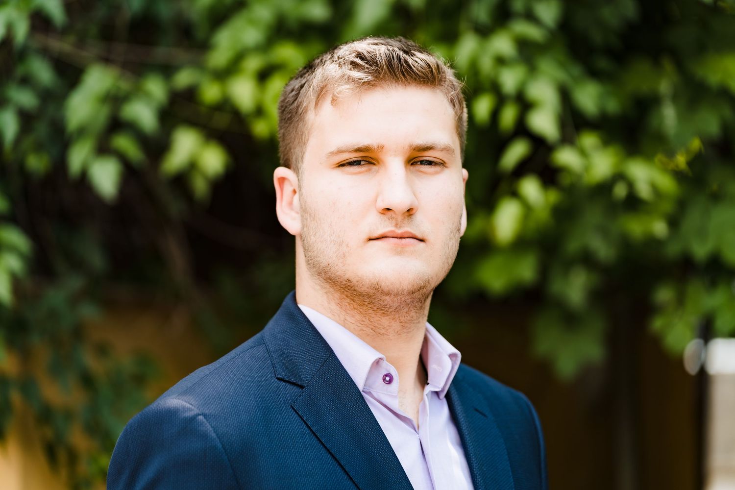 A young man with short light hair wears a navy blazer over a lavender collared shirt and stands before lush green foliage.