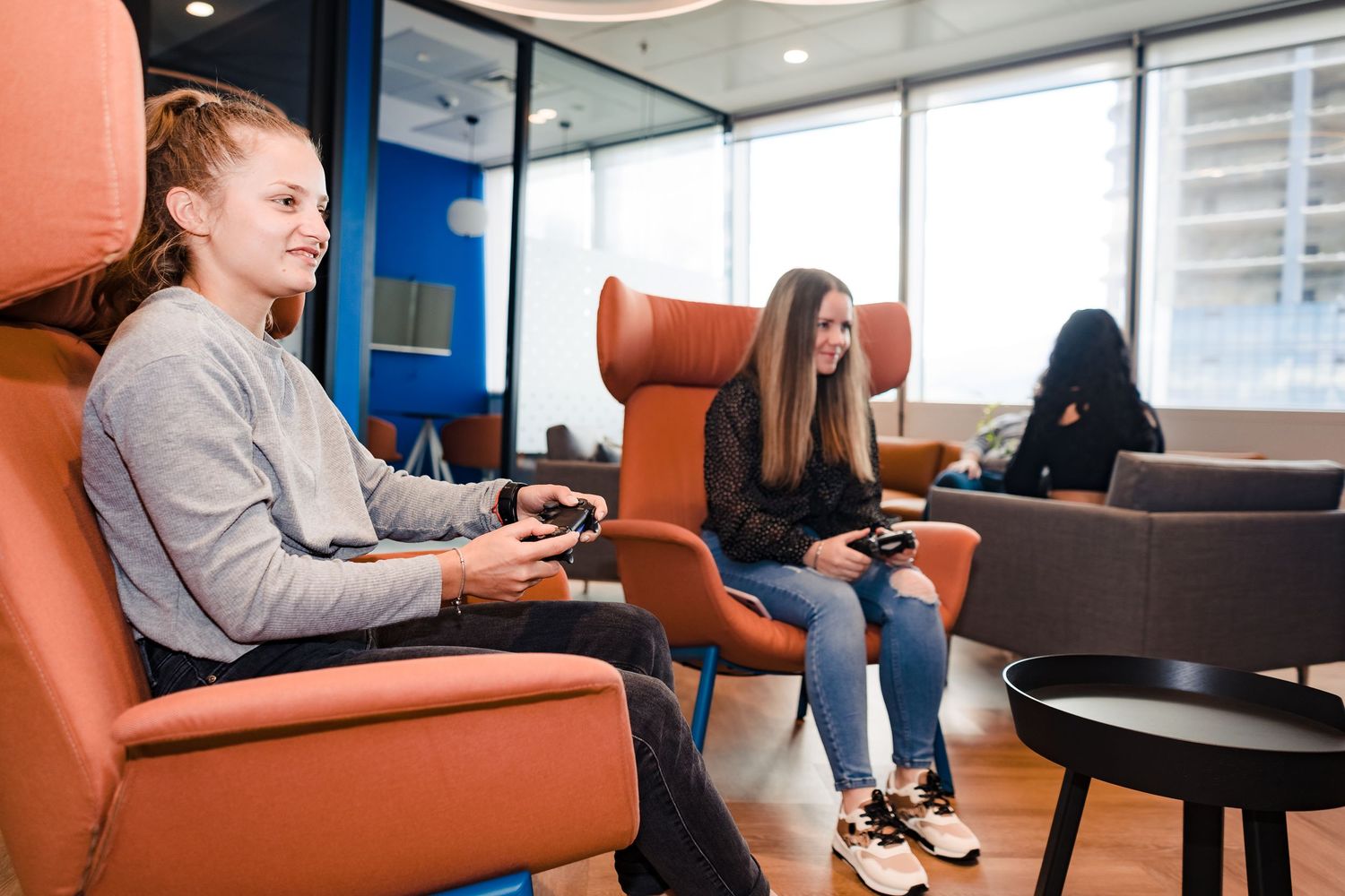 Two women sit in orange armchairs playing video games with controllers in a sunny office lounge with large windows.