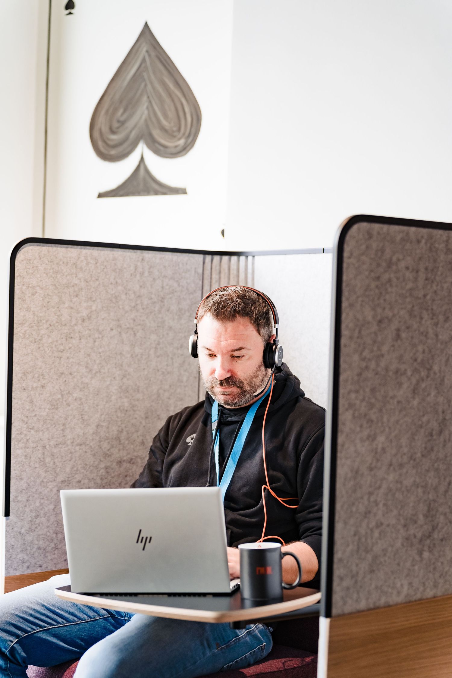 Man with headphones and a lanyard works on an HP laptop in a felt-paneled office booth with a spade symbol on the wall.
