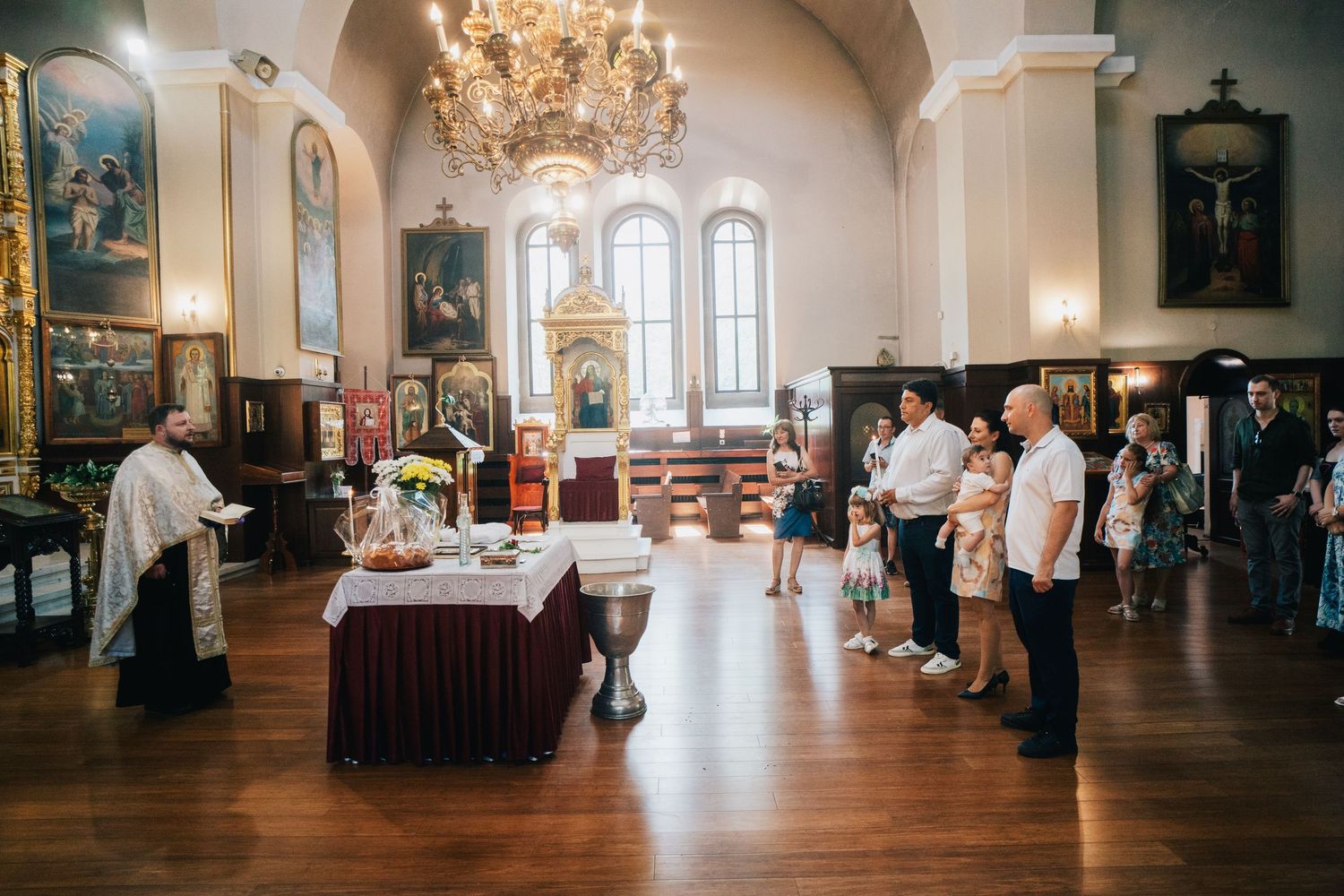 Family members stand in a bright Orthodox church with a chandelier and icons while a priest prepares for a baby's baptism.