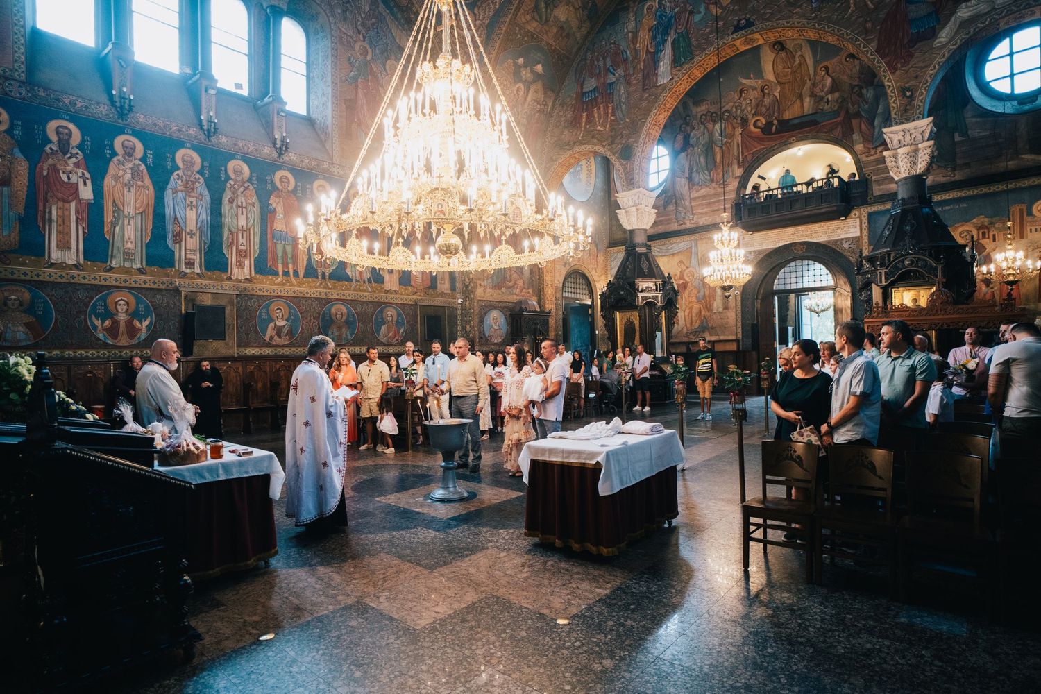 Priest in white vestments leads a ceremony inside an ornate church with a large chandelier, painted icons and gathered attendees.