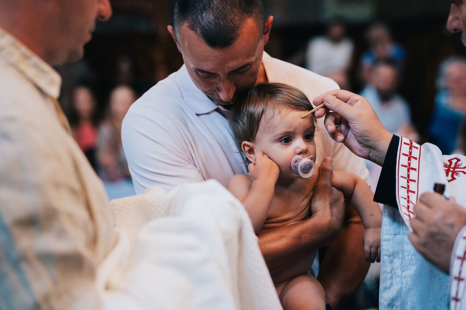 A baby with a pacifier is held by a man while a priest anoints the child's forehead during a traditional Orthodox christening ceremony.