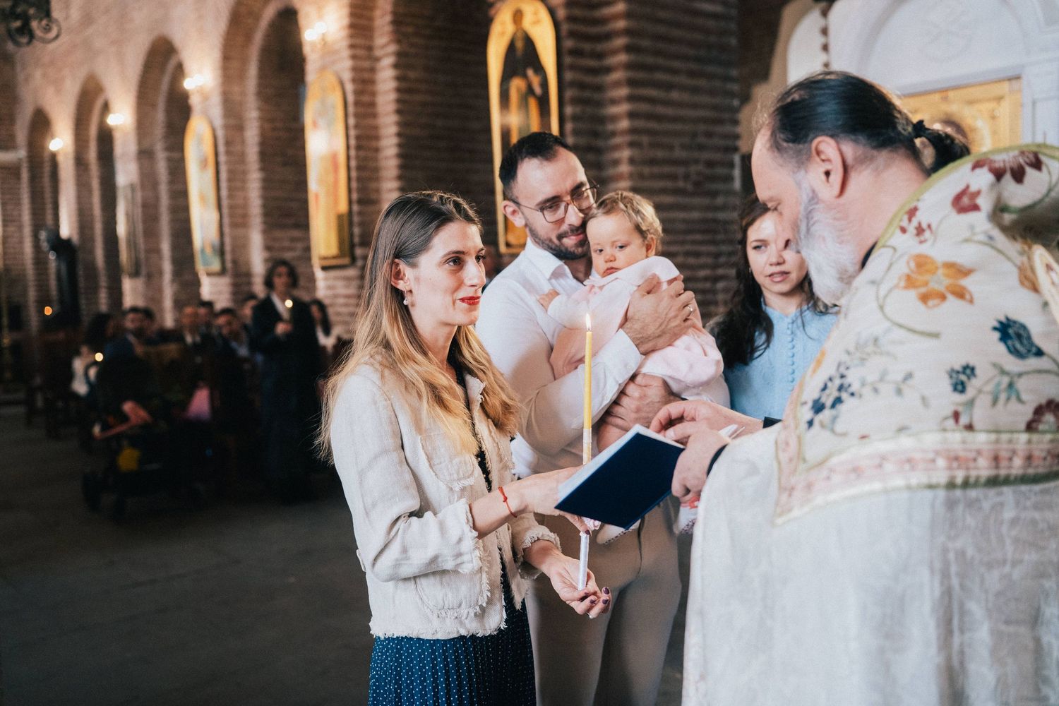 Parents hold a baby during an Orthodox christening as a priest in ornate vestments reads from a book inside a church.