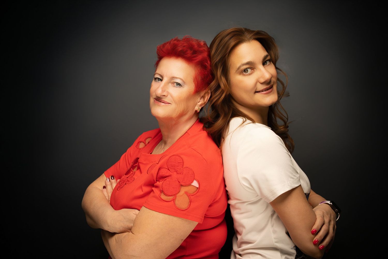 Two women stand back to back with arms crossed in a studio portrait, one in a red top with red hair, the other in white.