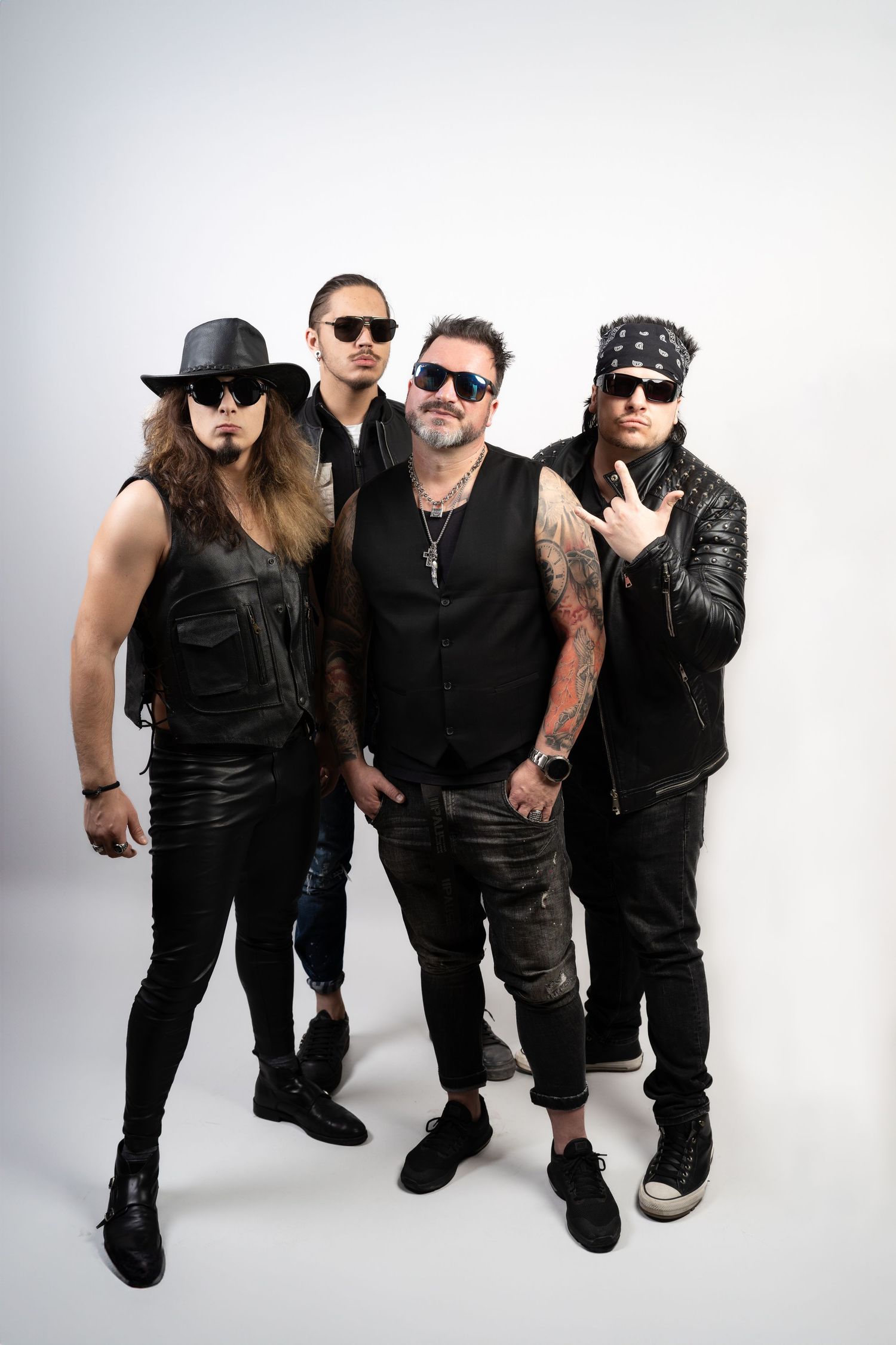 Rock band in black leather, sunglasses, and bandana pose confidently together on a white studio background, one flashing a hand sign.