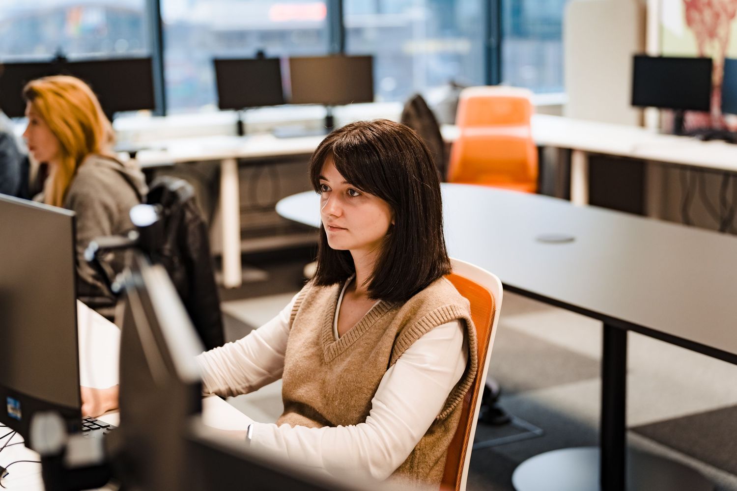 A woman with dark hair sits at a desk working on a computer in a bright modern office while a colleague works in the background.