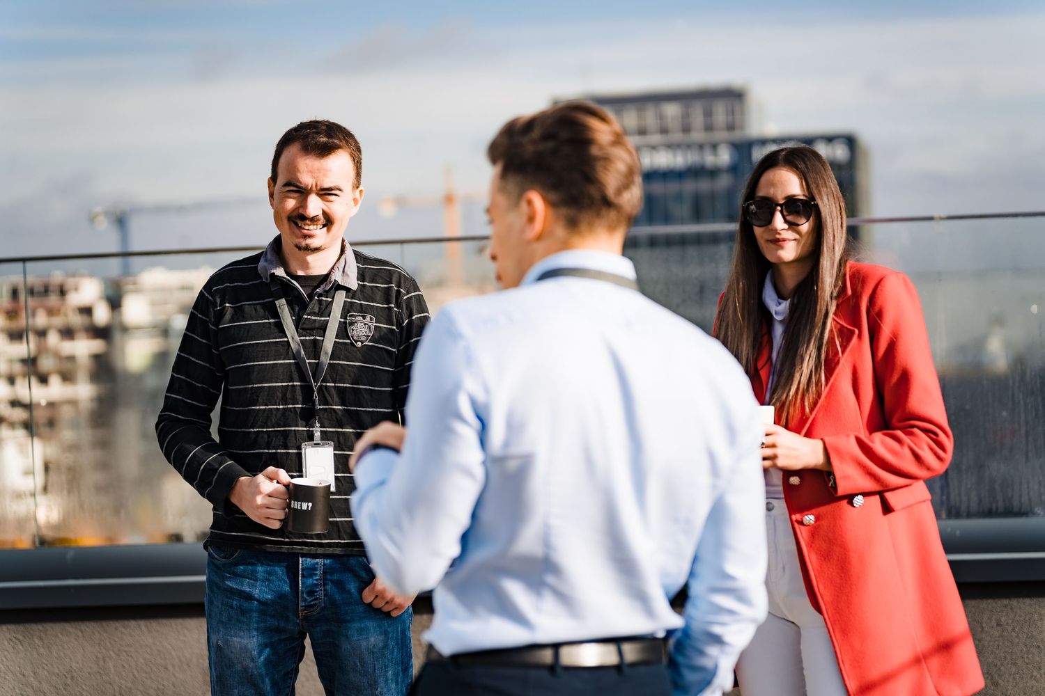 Three coworkers with coffee mugs chat on a rooftop terrace overlooking the city during a casual work break.