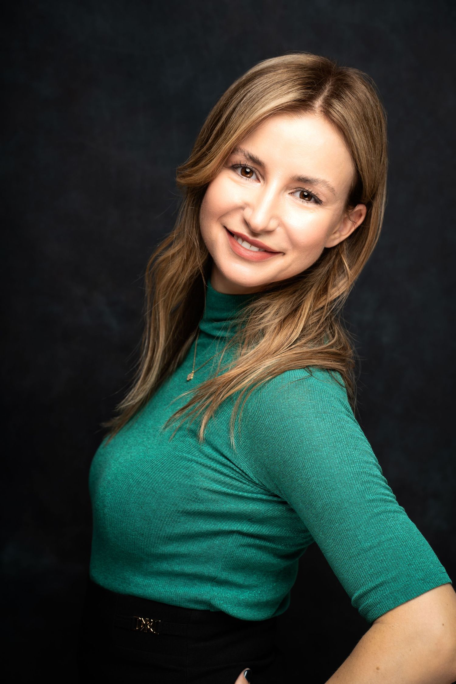 A smiling woman with long blonde hair wears a green knit turtleneck top and poses against a dark studio background.