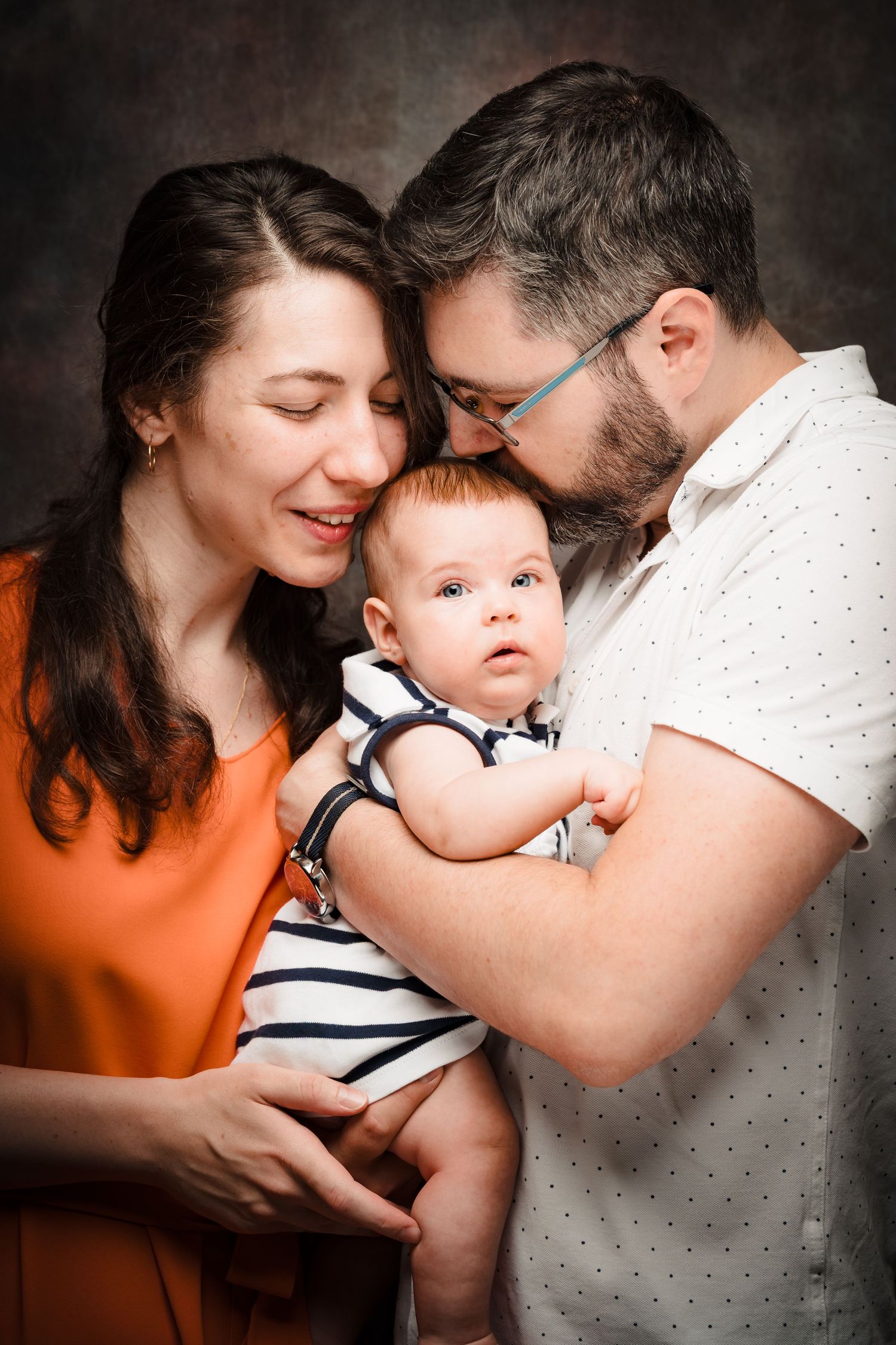 A mother and bearded father in glasses tenderly kiss their baby's head, wearing orange and white, in an intimate studio family portrait.