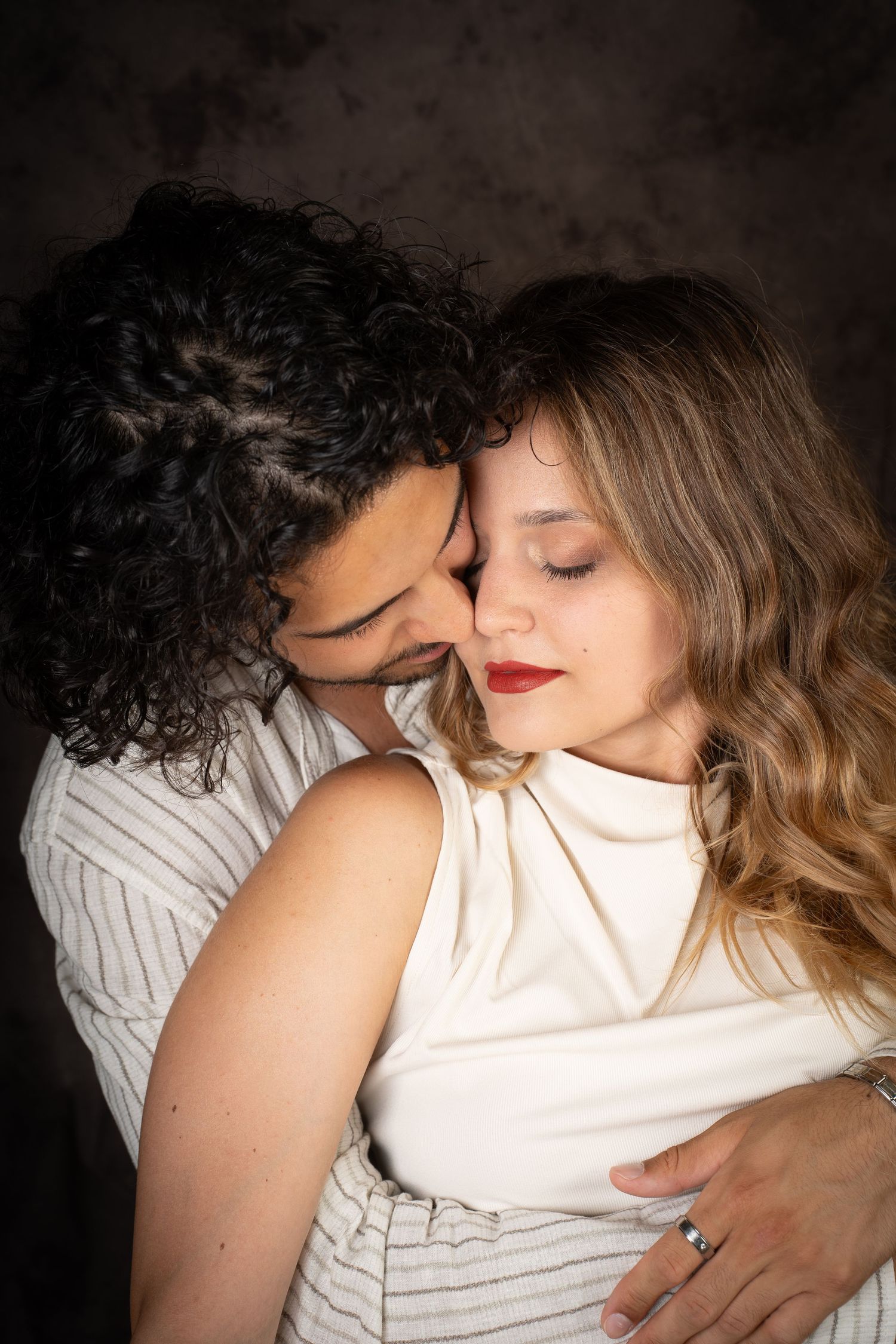 Romantic studio portrait of a curly-haired man embracing a smiling woman with red lipstick and wavy blonde hair from behind, eyes closed.