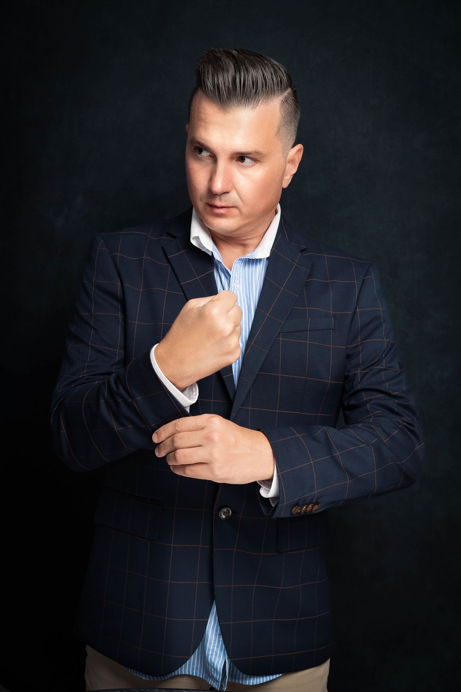 Stylish man in a navy checked blazer and blue striped shirt adjusts his cuff in a moody studio portrait.