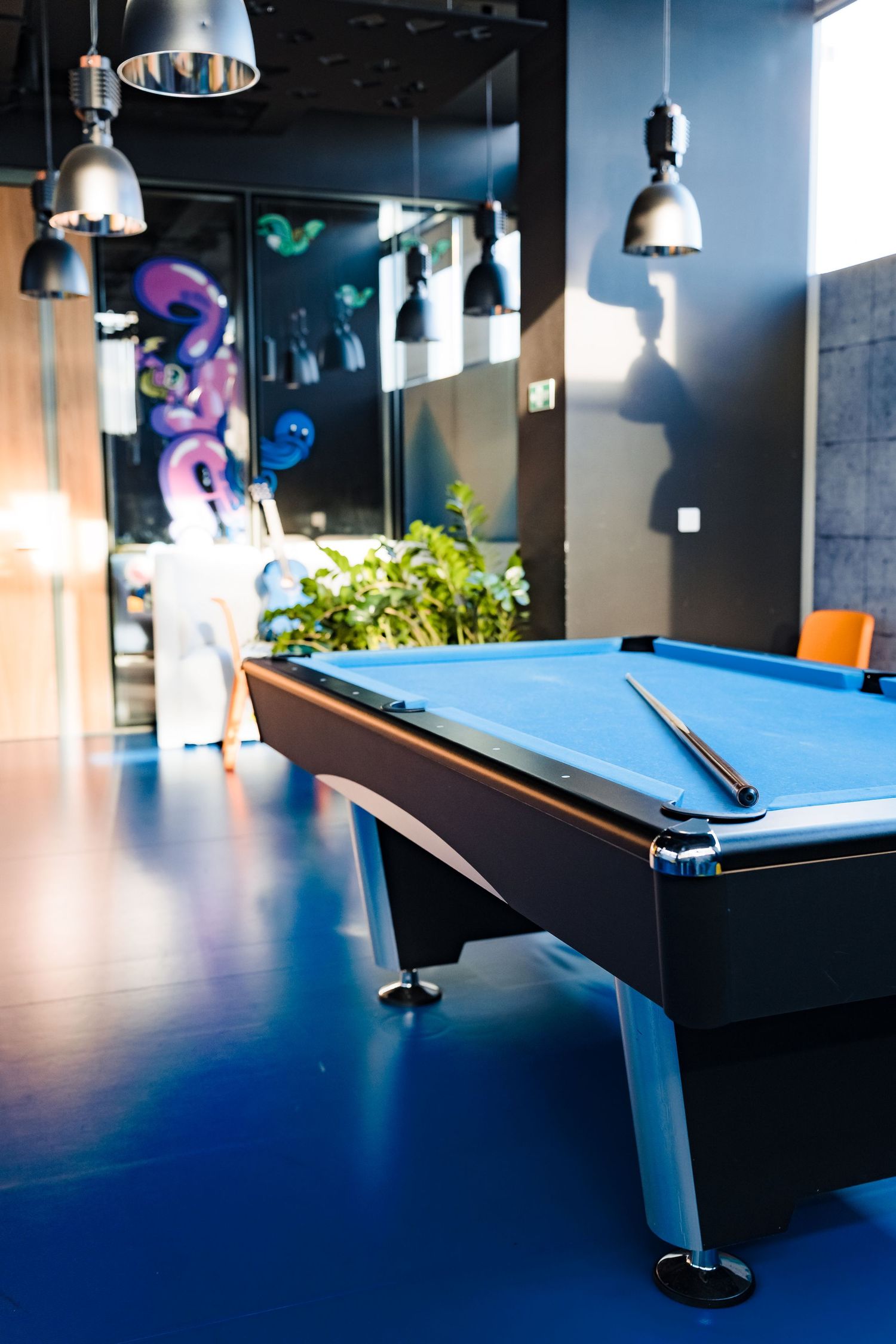 Blue pool table with a cue stick in a modern game room with pendant lamps, colorful graffiti art and a green plant.