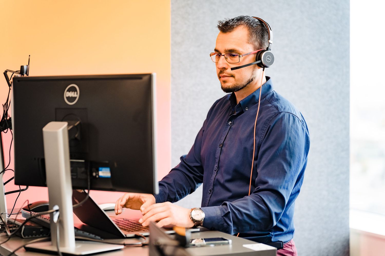 Man with glasses and a headset works at a standing desk in front of a Dell monitor in a bright modern office.