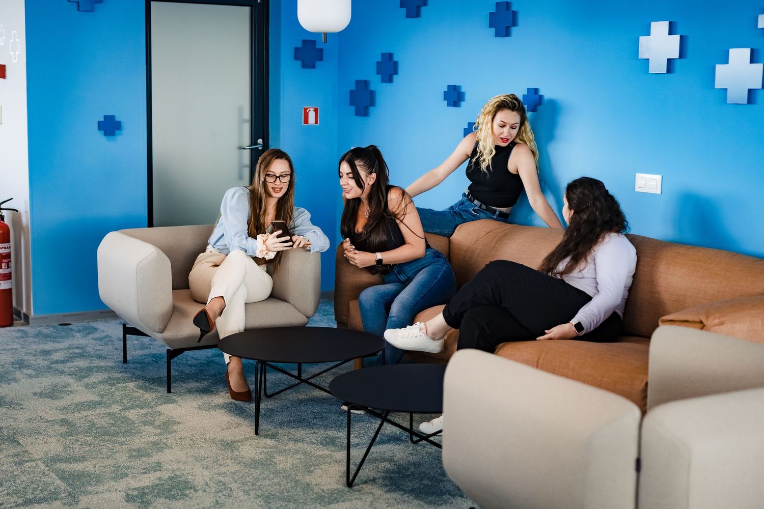 Four women socialize in a modern blue office lounge with beige and tan sofas, black coffee tables and cross-shaped wall decor.