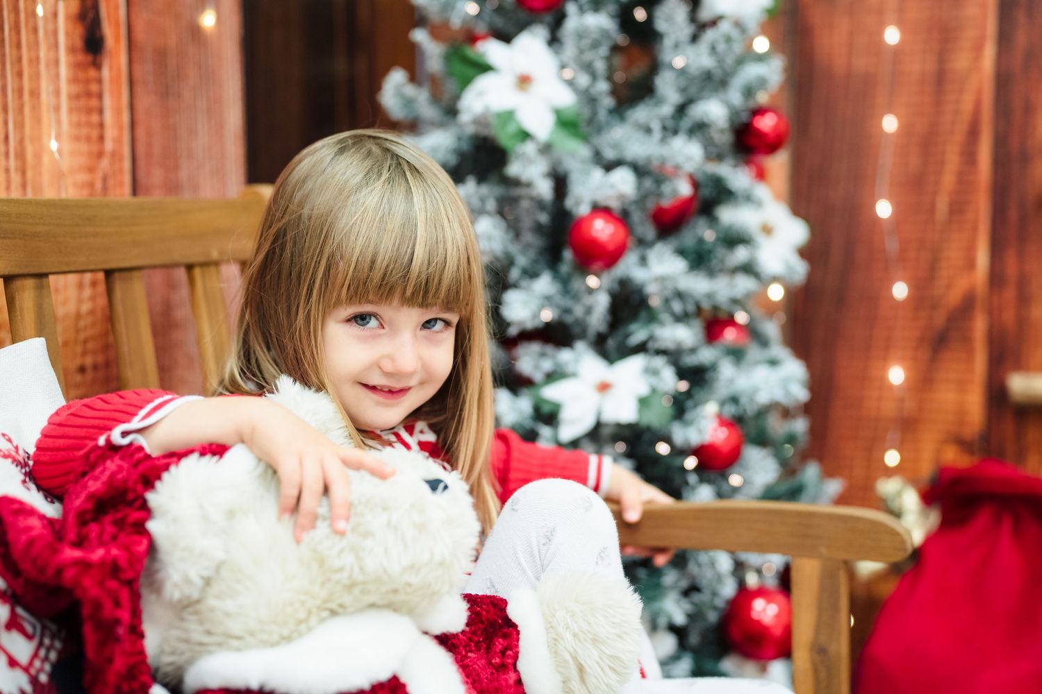 Happy girl hugs a plush white fur toy near a decorated Christmas tree with red ornaments and warm holiday lights.