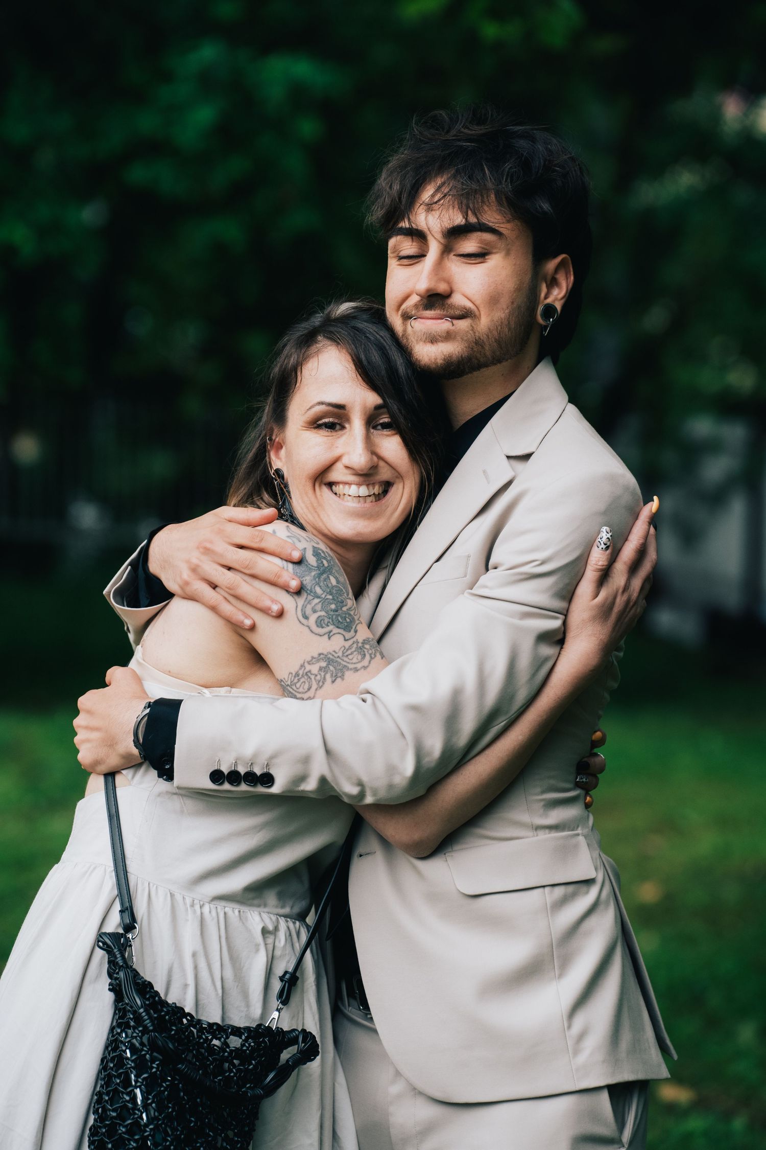A smiling woman with a tattooed arm hugs a bearded man in a beige suit outdoors in front of lush green foliage.