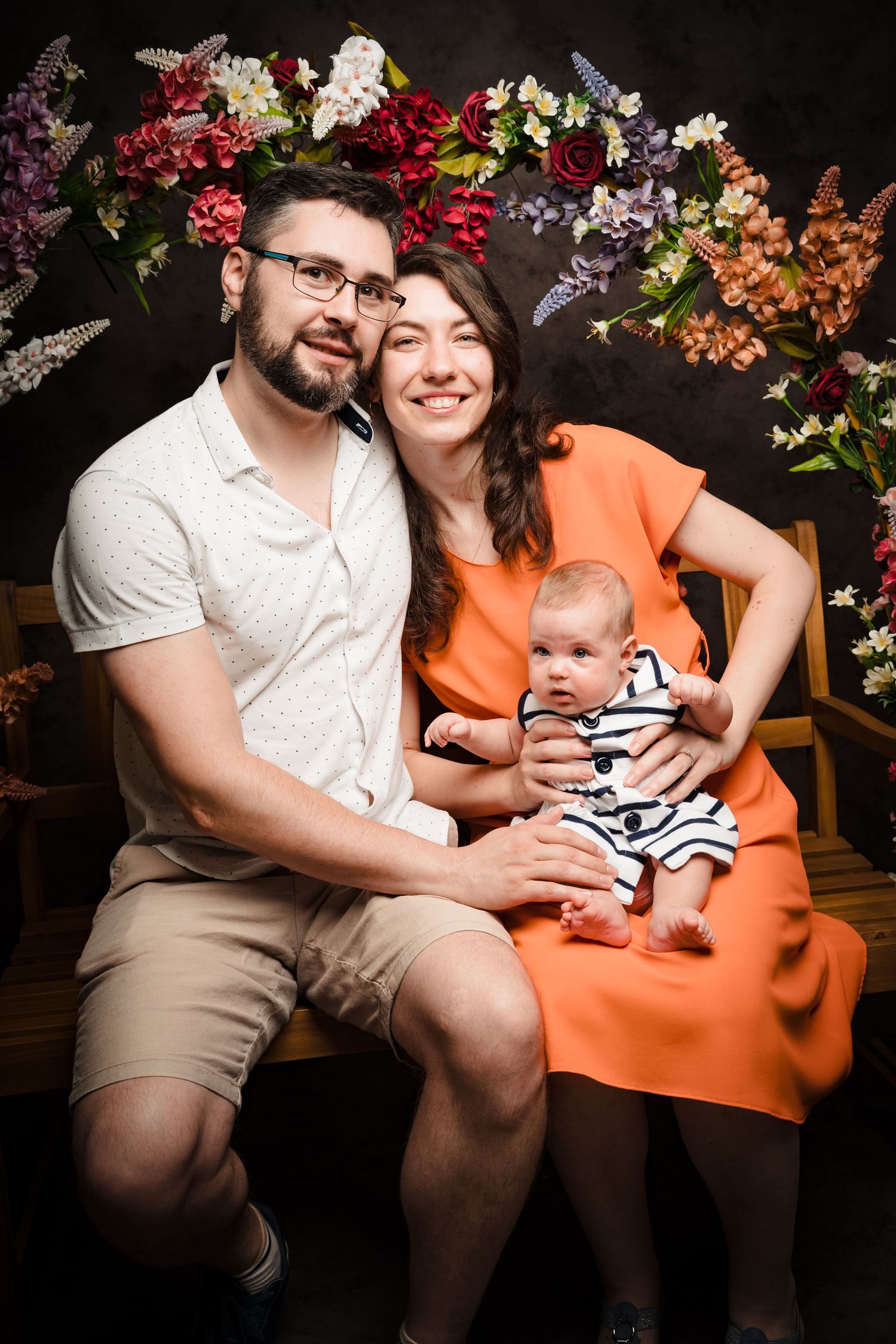 A happy family of three sits on a wooden bench with their baby, framed by colorful flower arrangements in a studio portrait.
