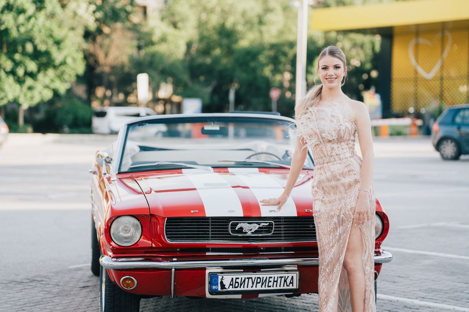 Woman in a long sparkling blush gown with a high slit stands beside a red Ford Mustang convertible with white racing stripes.