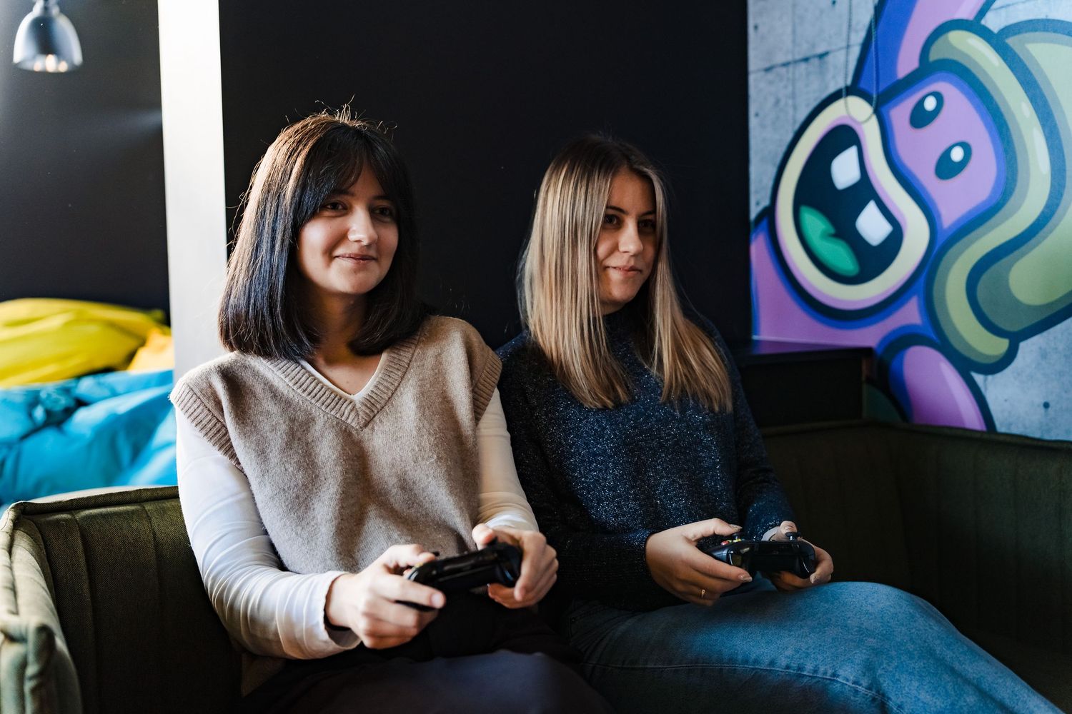 Two young women sit on a couch playing video games with controllers in front of a colorful graffiti wall.