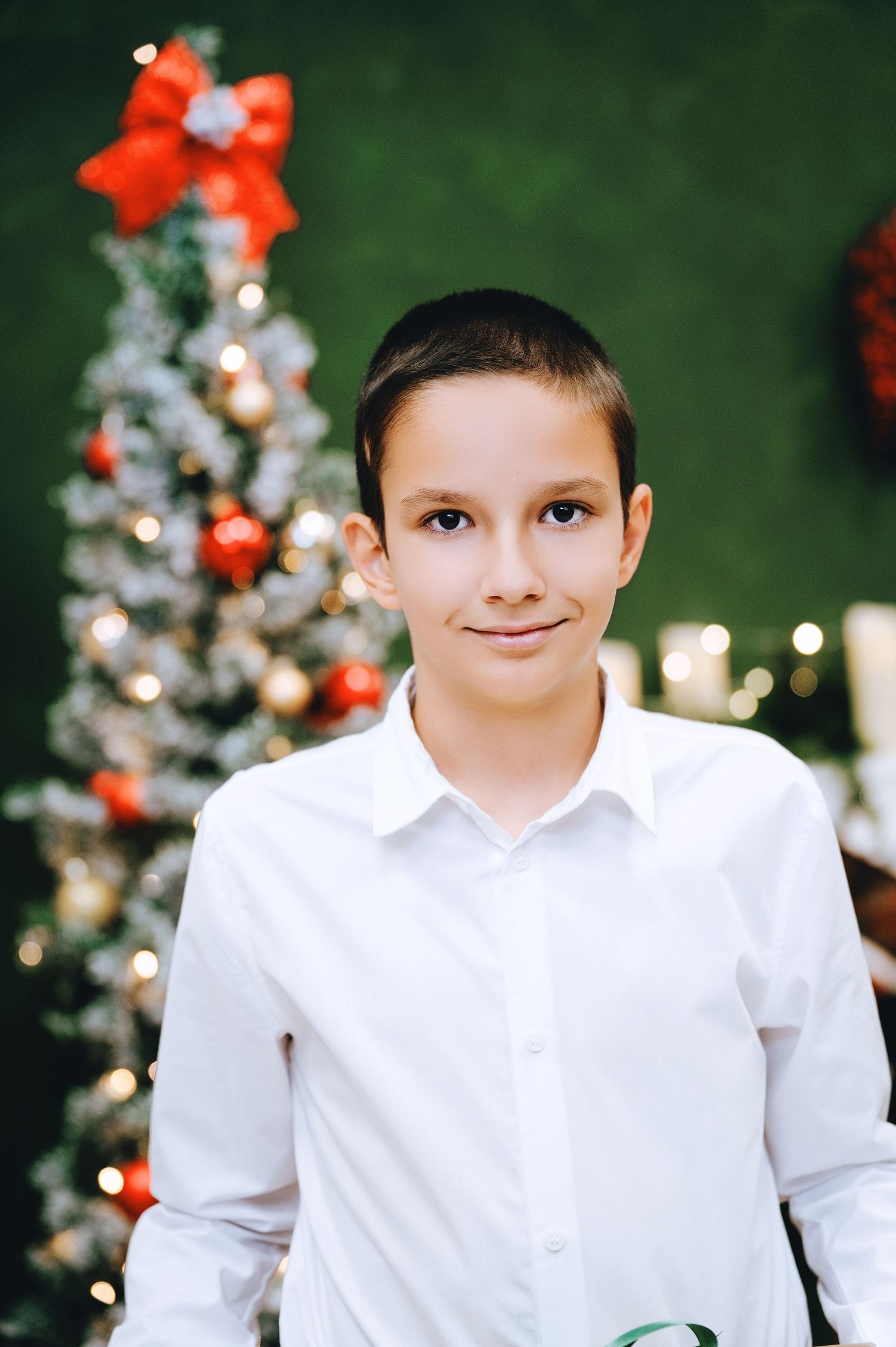 Smiling boy in a white button-up shirt stands in front of a decorated Christmas tree with red bows and warm string lights.