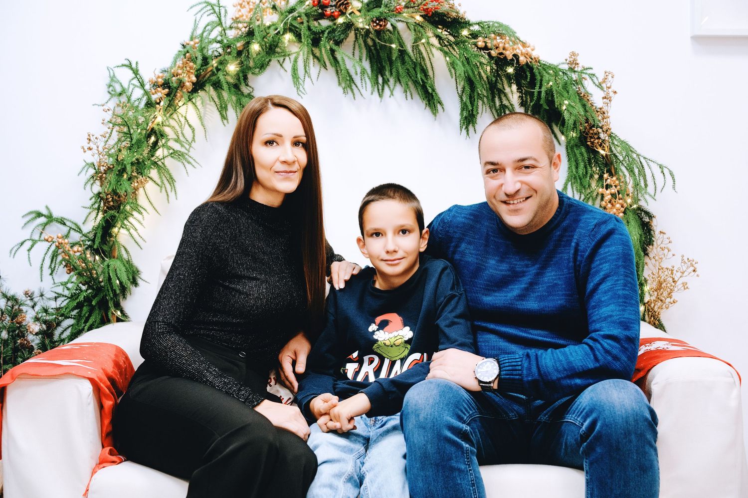 Smiling family of three, a woman, a man and a young boy, sitting together on a white sofa in front of a green floral wreath arch.
